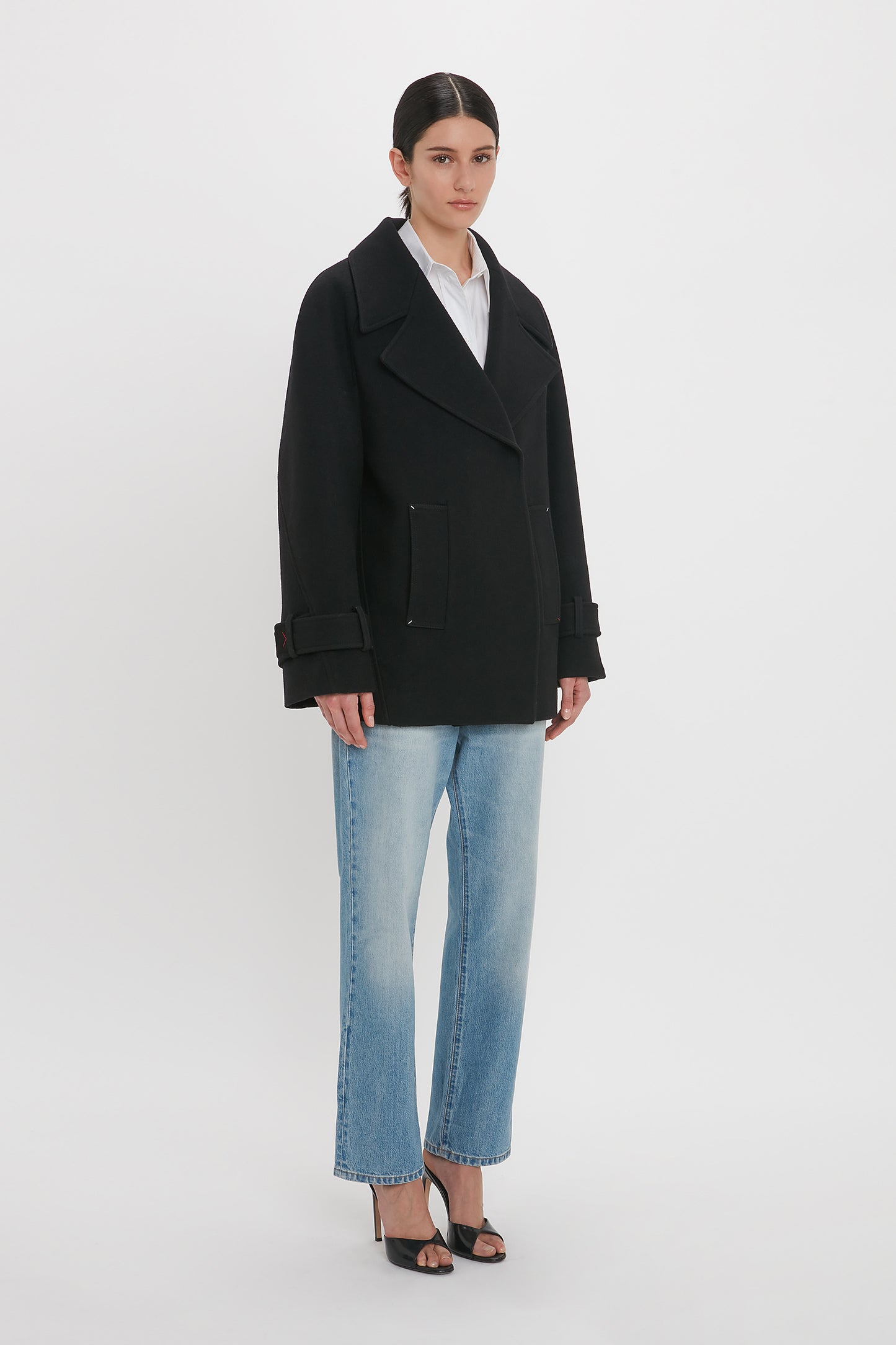 A person stands against a white background wearing an Oversized Pea Coat In Black by Victoria Beckham, light blue jeans, and black heeled sandals.