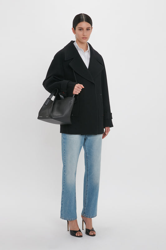 A person stands against a white background wearing an Oversized Pea Coat In Black by Victoria Beckham, light blue jeans, black high-heeled sandals, and holding a black handbag.