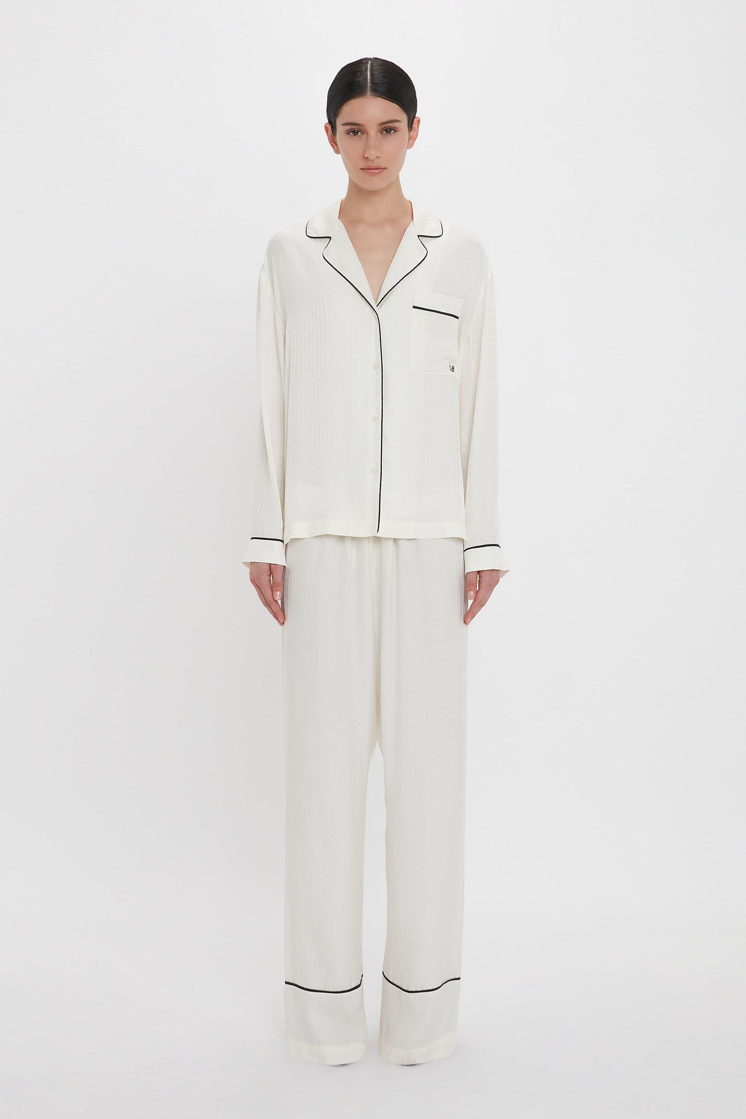 Person standing against a plain background wearing the VB Monogram Pyjama Sleep Set In Ivory by Victoria Beckham. The pajama has a collar, pocket, and buttons down the front. The pants are loose-fitting, making it an ideal loungewear set for ultimate comfort.