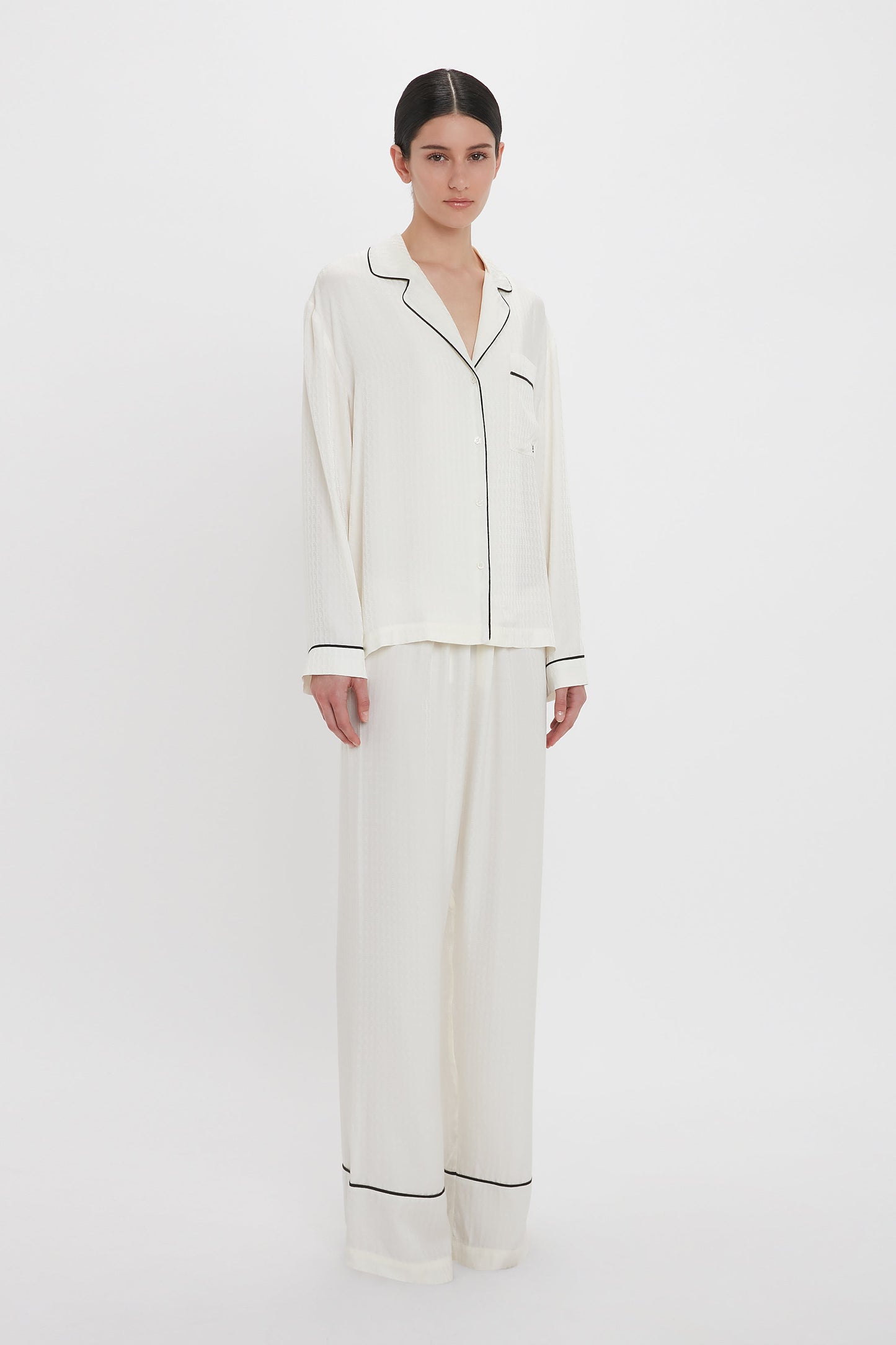 A person stands against a white background, wearing the Victoria Beckham VB Monogram Pyjama Sleep Set In Ivory consisting of a white button-up pajama top and matching wide-leg pajama pants with black trim on the cuffs and collar.