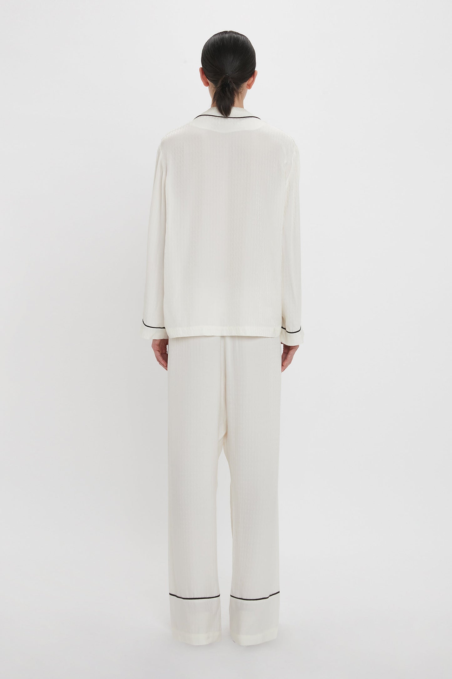 A person stands facing away from the camera, wearing a white VB Monogram Pyjama Sleep Set In Ivory with black trim by Victoria Beckham, in a minimalistic, well-lit background.