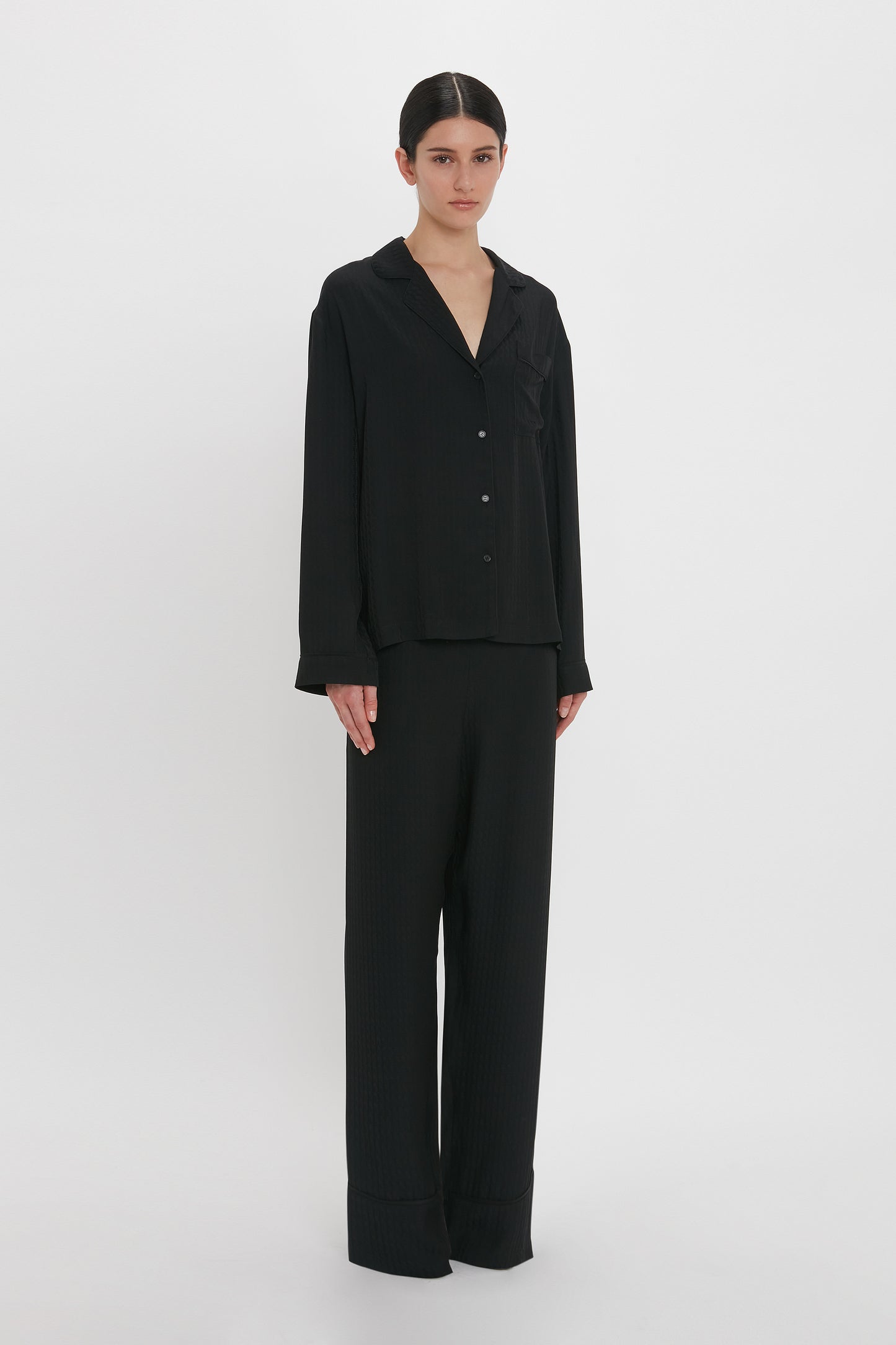 A person stands against a plain white background wearing the VB Monogram Pyjama Sleep Set In Black by Victoria Beckham.