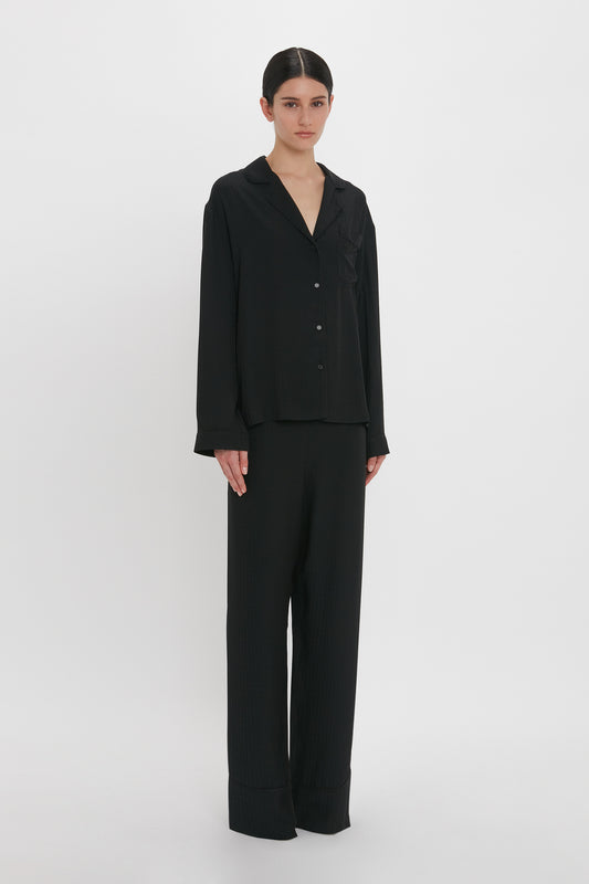 A person stands against a plain white background wearing the VB Monogram Pyjama Sleep Set In Black by Victoria Beckham.