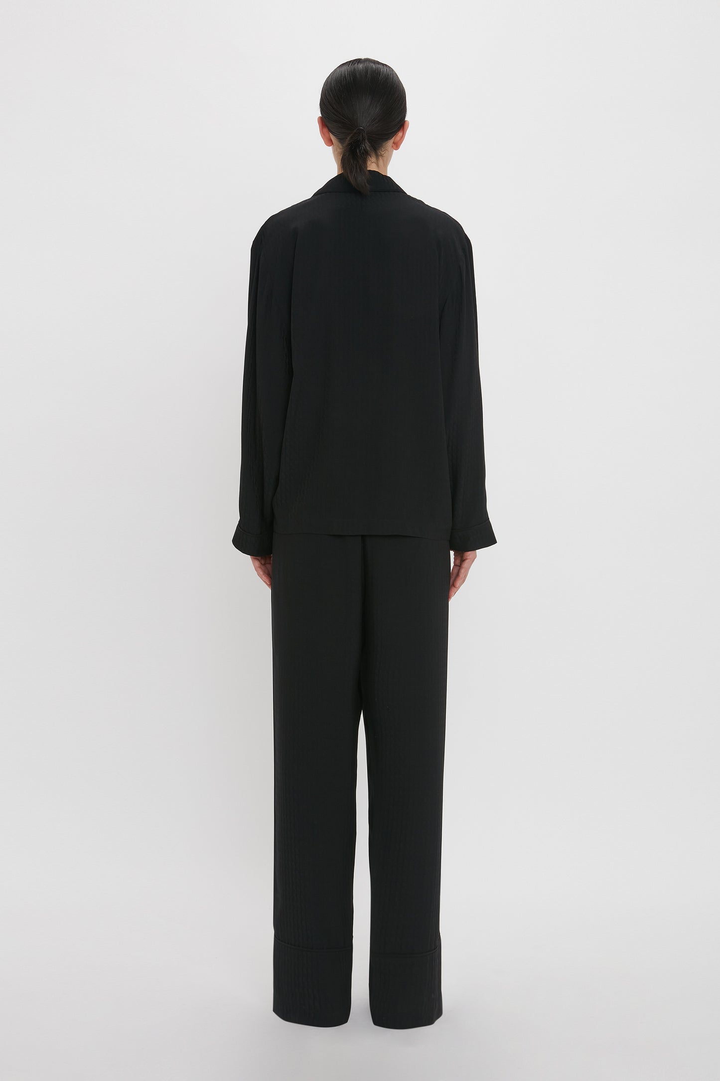 Back view of a person with dark hair tied back, standing against a plain white background, wearing a loose-fitting black long-sleeved top and black pants that resemble the VB Monogram Pyjama Sleep Set In Black by Victoria Beckham.