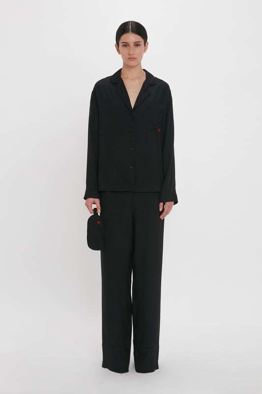 A person is standing against a plain white background, wearing a matching black VB Monogram Pyjama Sleep Set In Black made of 100% silk by Victoria Beckham. They hold a small black object in one hand.