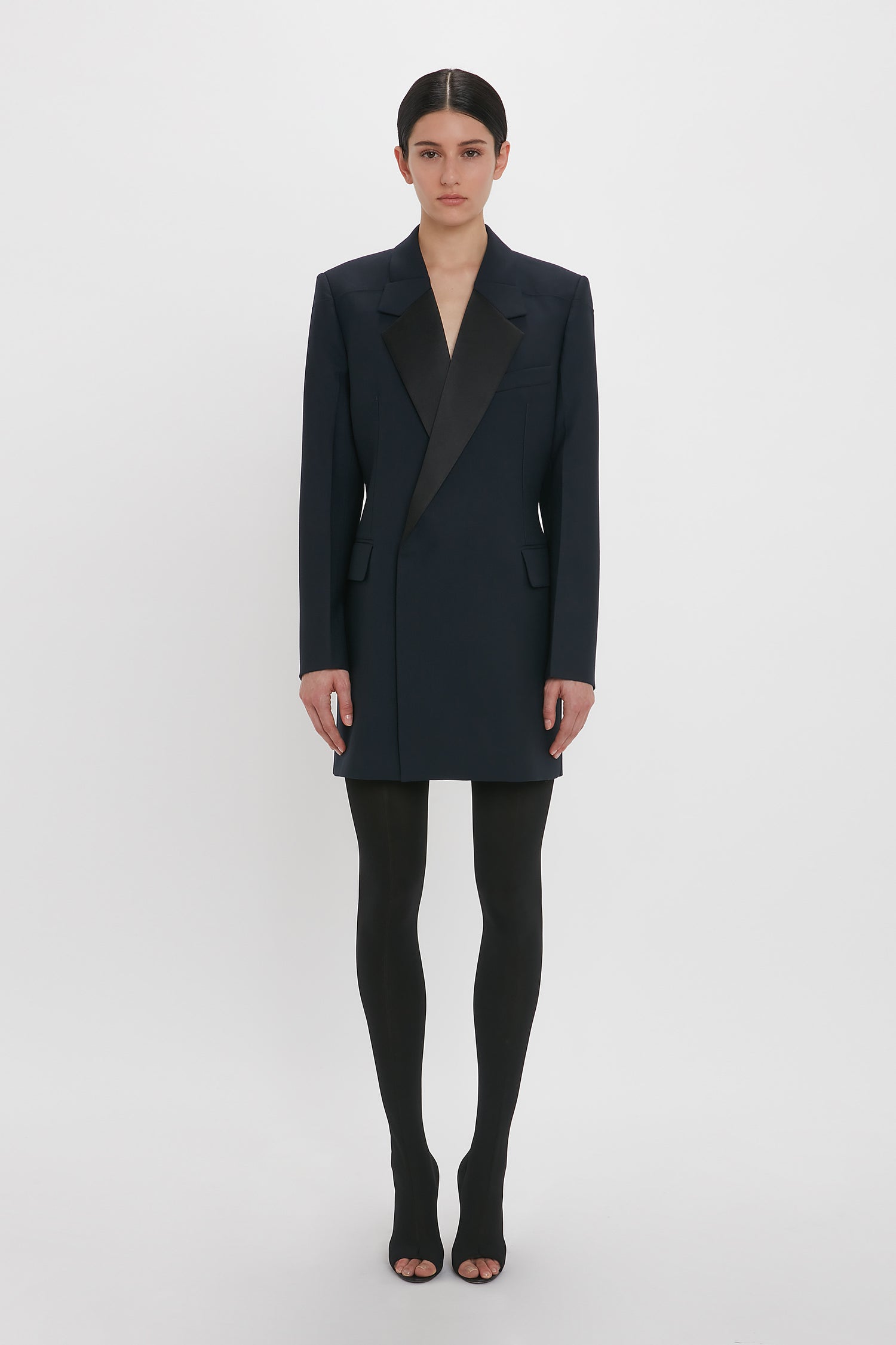 A person stands against a plain background, wearing a black Exclusive Fold Shoulder Detail Dress In Midnight reminiscent of Victoria Beckham's style and black tights. Their hands are relaxed at their sides.