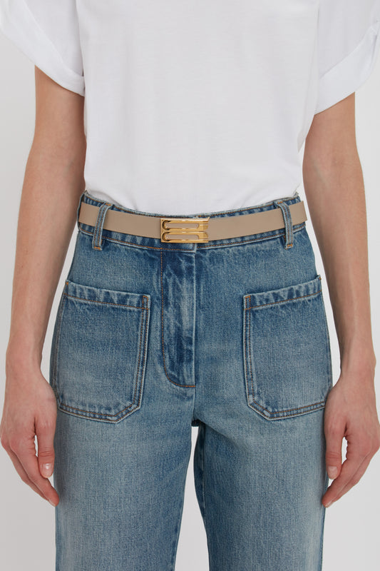 A fashion-forward individual is wearing a white T-shirt and blue jeans with large pockets, secured by an Exclusive Frame Belt In Beige Leather featuring a gold buckle from Victoria Beckham.