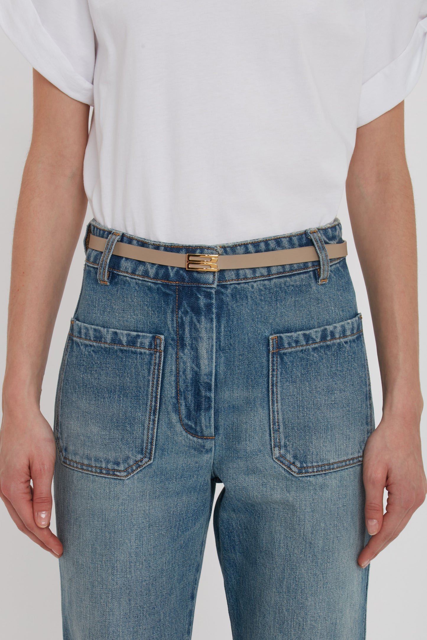 A person in a white t-shirt and blue jeans with large pockets wears a beige Exclusive Micro Frame Belt In Beige Leather by Victoria Beckham.