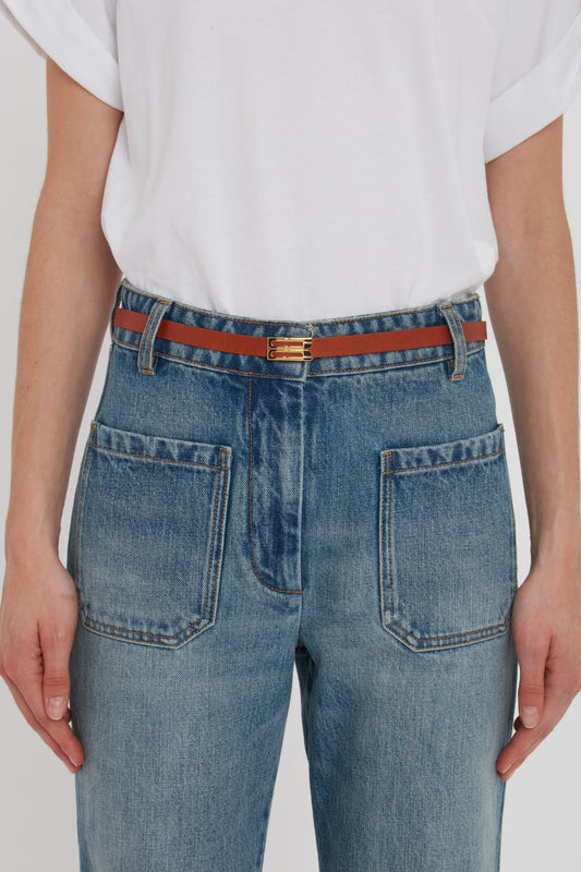 Person wearing a white shirt tucked into blue high-waisted jeans with two front pockets and a thin brown Exclusive Micro Frame Belt In Tan Leather by Victoria Beckham featuring gold hardware.