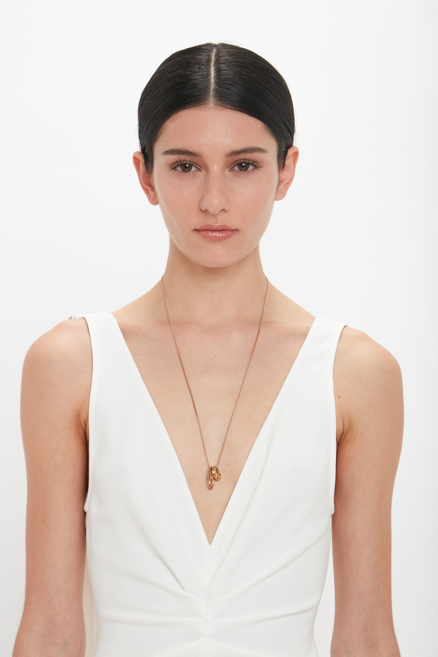 A woman with straight black hair, wearing a white sleeveless V-neck dress and an Exclusive Abstract Charm Necklace In Light Gold by Victoria Beckham, stands against a plain white background.