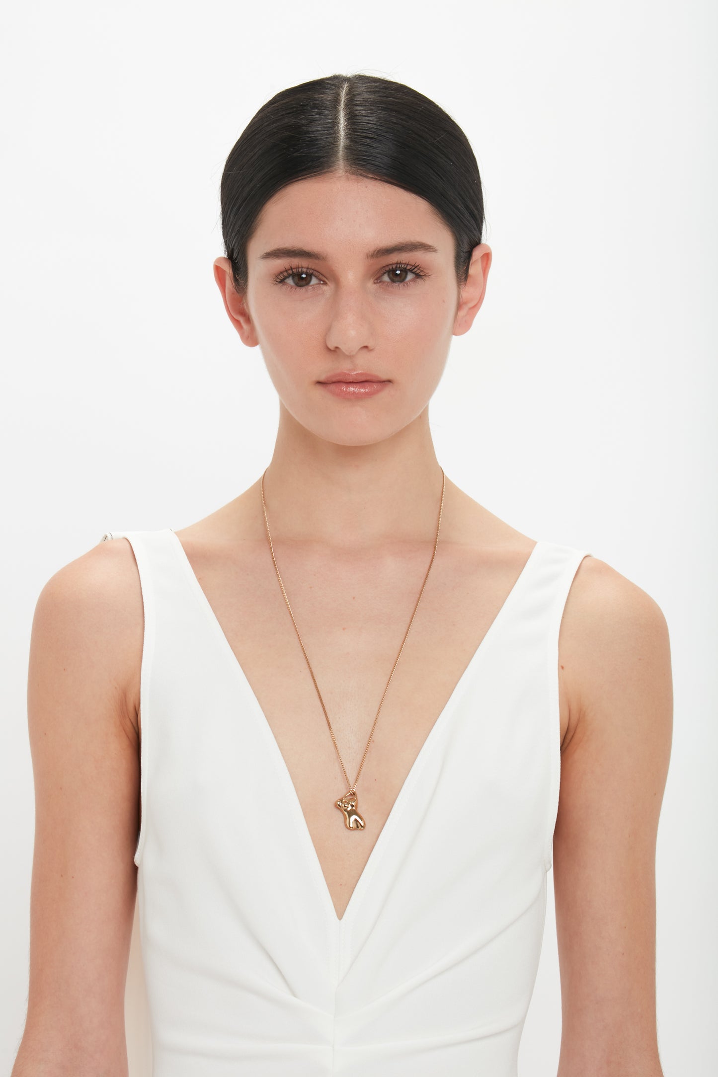 A woman with straight dark hair is wearing a white, deep V-neck dress and a gold Exclusive Body Charm Necklace In Light Gold by Victoria Beckham with a heart and key pendant made from 100% brass. She is standing against a plain white background.