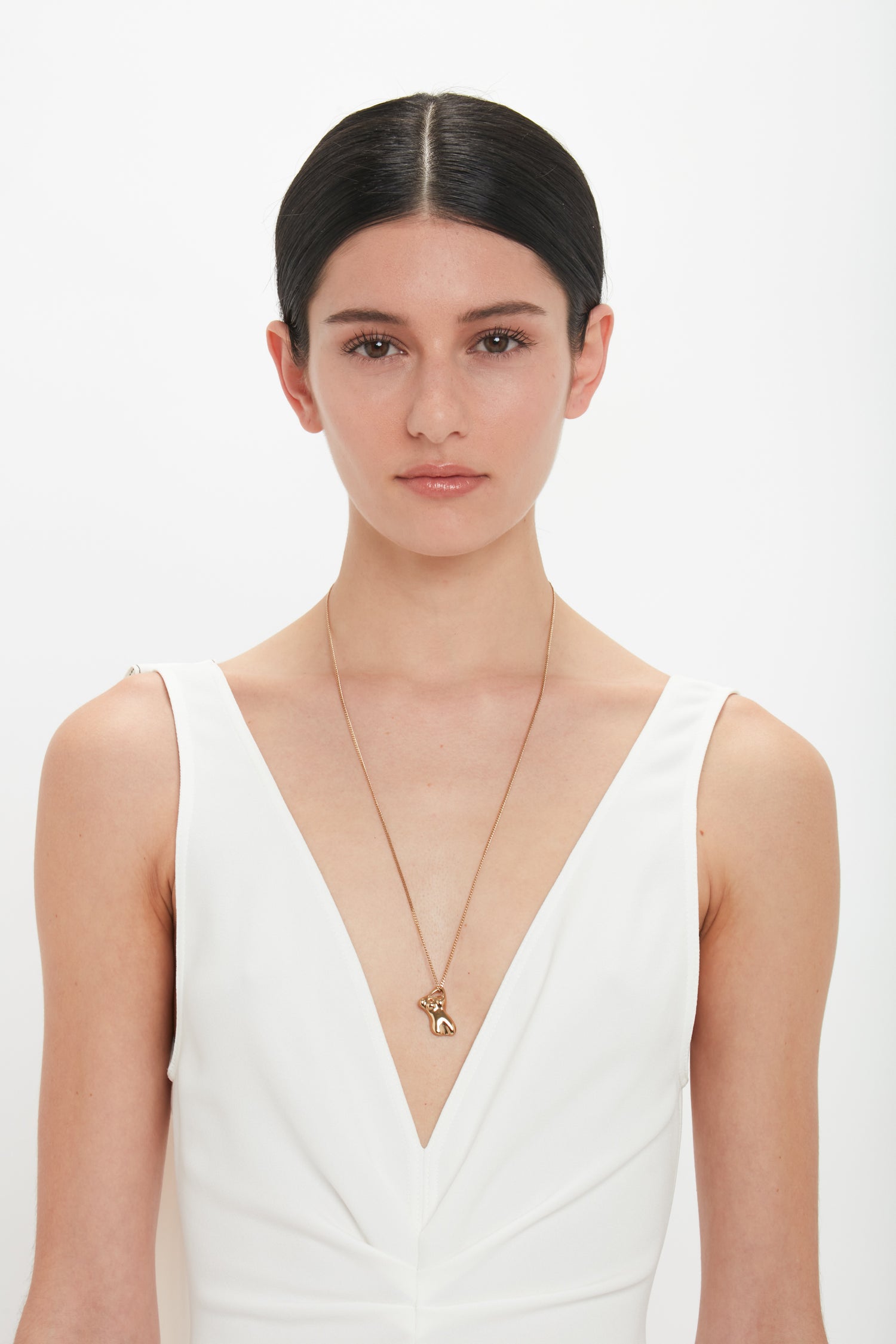 A woman with straight dark hair is wearing a white, deep V-neck dress and a gold Exclusive Body Charm Necklace In Light Gold by Victoria Beckham with a heart and key pendant made from 100% brass. She is standing against a plain white background.