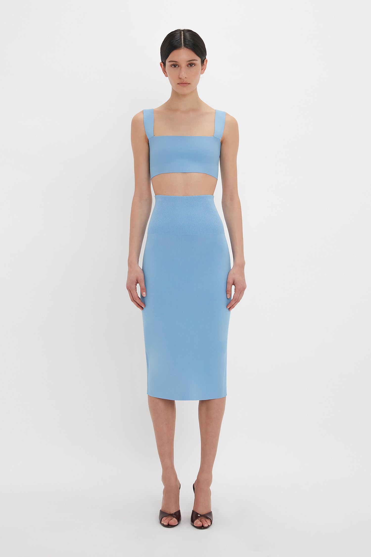 A person stands facing the camera, wearing a light blue, ultra form-fitting two-piece outfit consisting of a Victoria Beckham Strap Bandeau Top In Marina and a fitted midi skirt. They are also wearing brown open-toe high heel sandals.