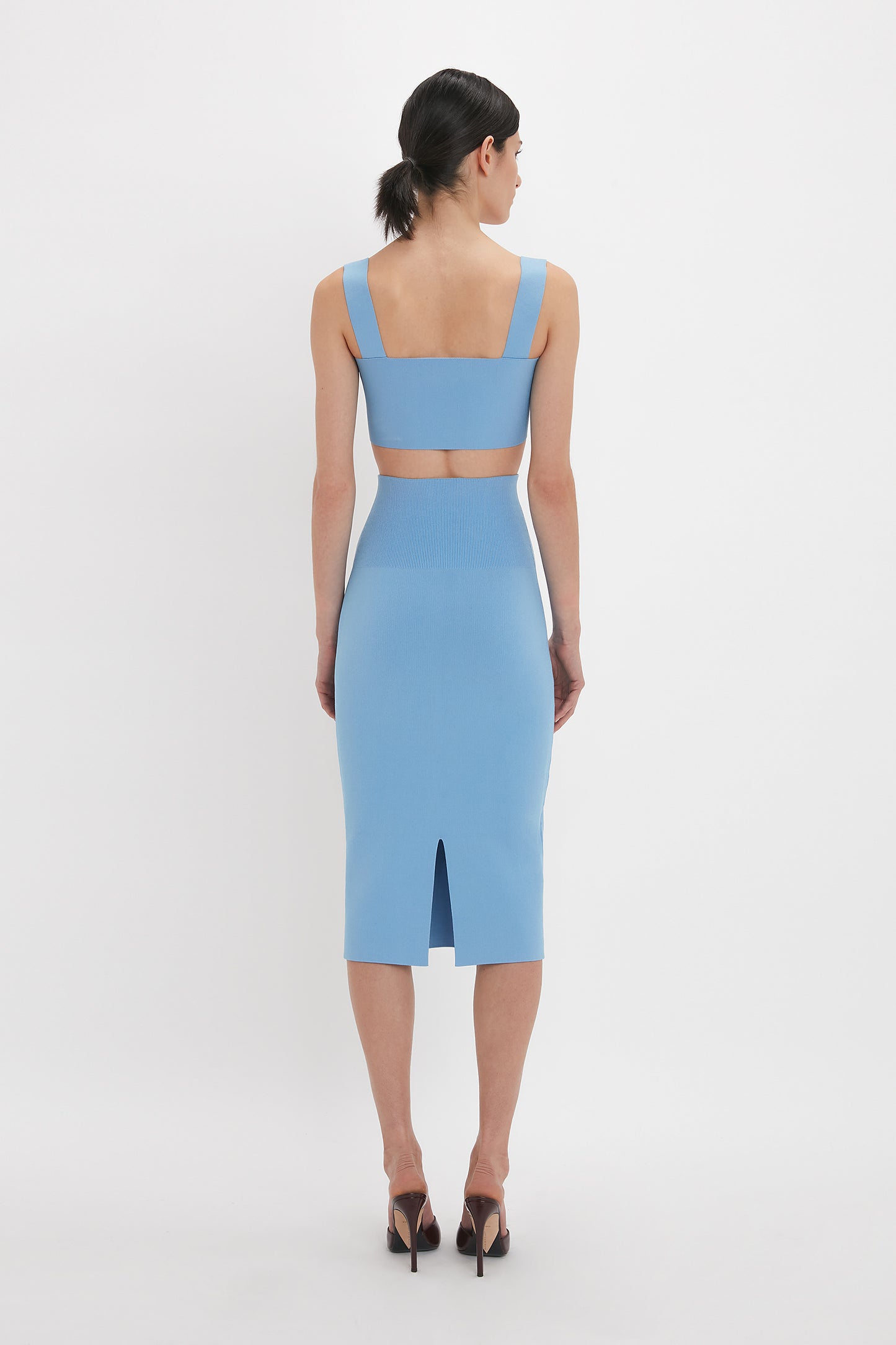 A person stands facing away, wearing a Strap Bandeau Top In Marina by Victoria Beckham with a cutout back and high heels, accentuating an ultra form-fitting silhouette.