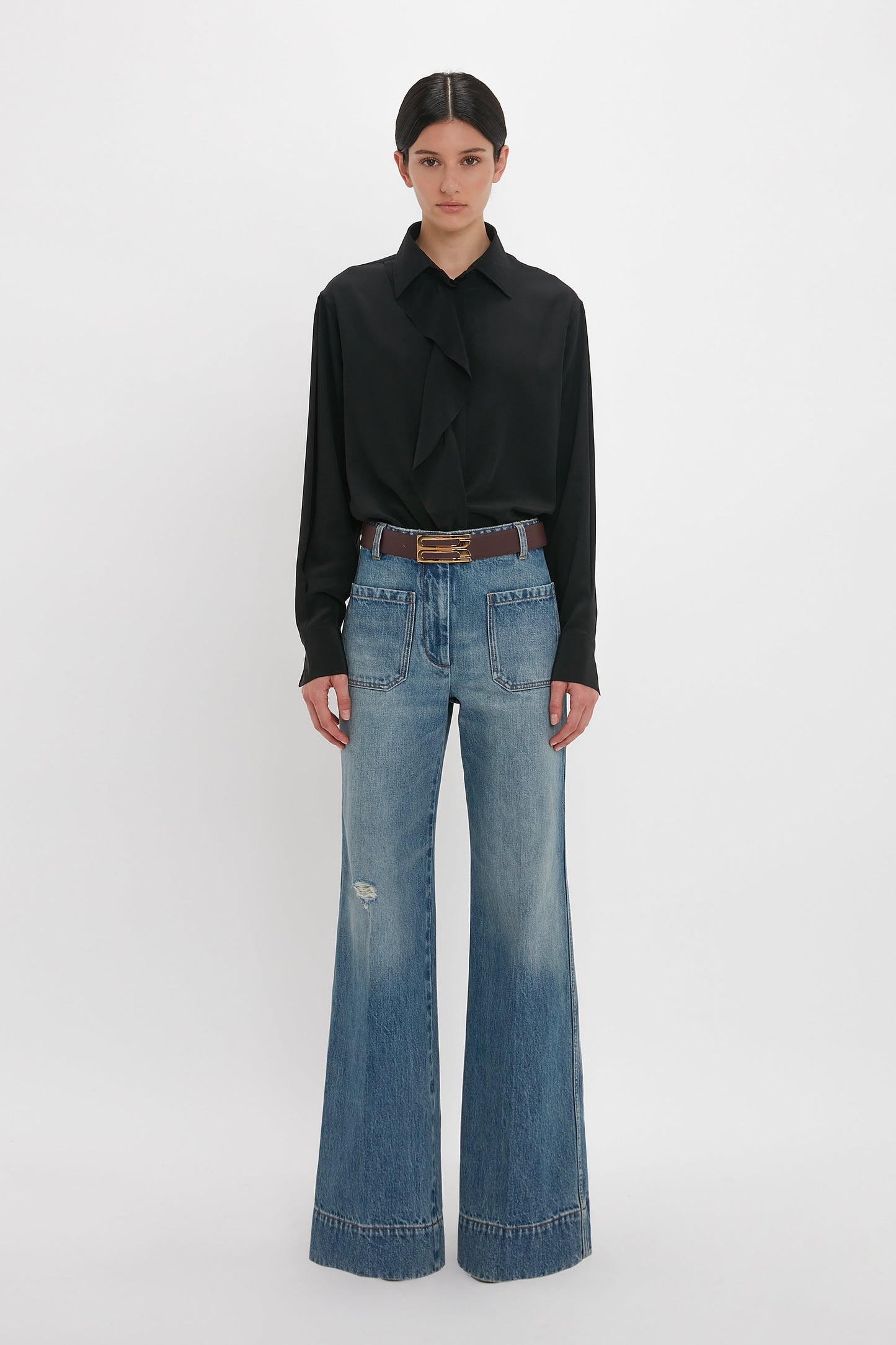 A woman stands against a white background wearing a Victoria Beckham black asymmetric ruffle blouse and distressed wide-leg jeans, accessorized with a brown belt.