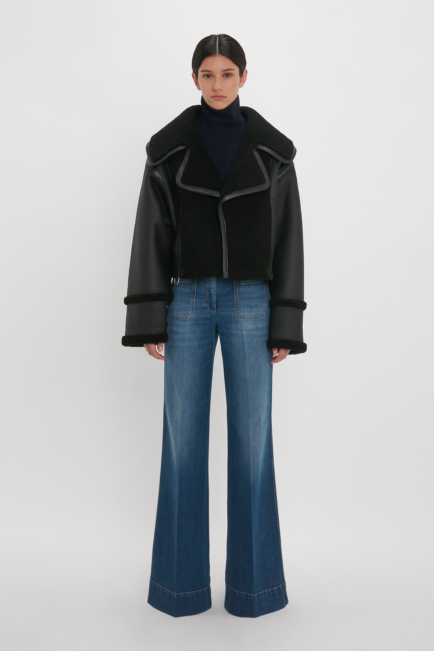 Person standing in front of a plain background, wearing the Victoria Beckham Shearling Jacket In Black over a dark turtleneck and wide-leg blue jeans for a modern classic look.