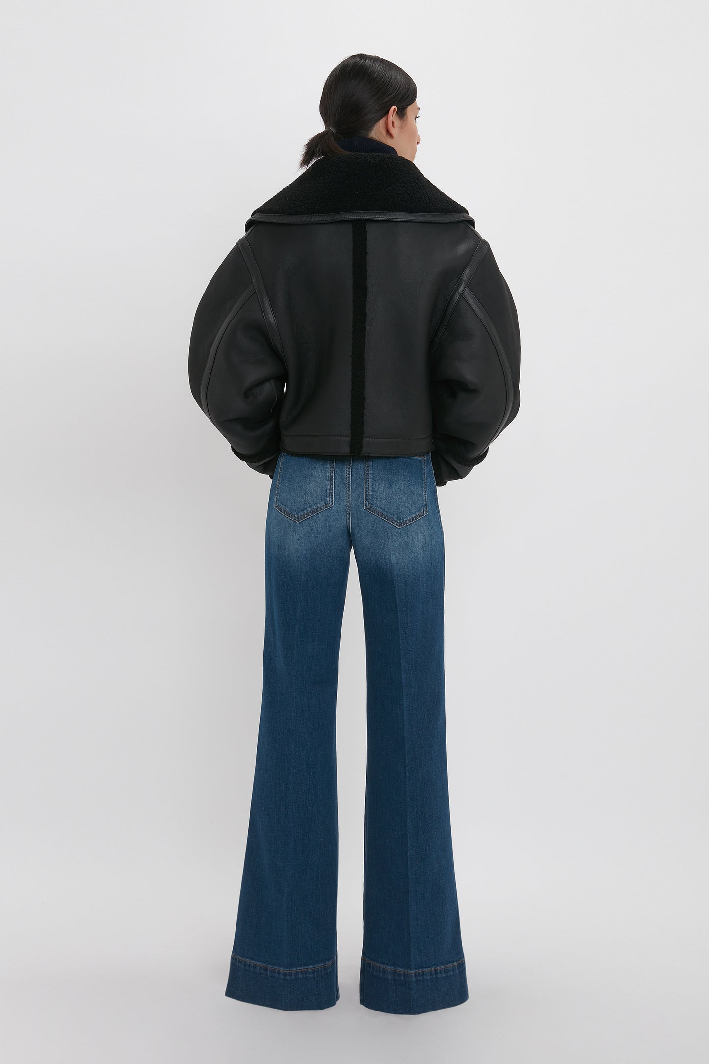 Person with long hair tied back, wearing a black Shearling Jacket In Black by Victoria Beckham and blue wide-leg jeans, standing and facing away.