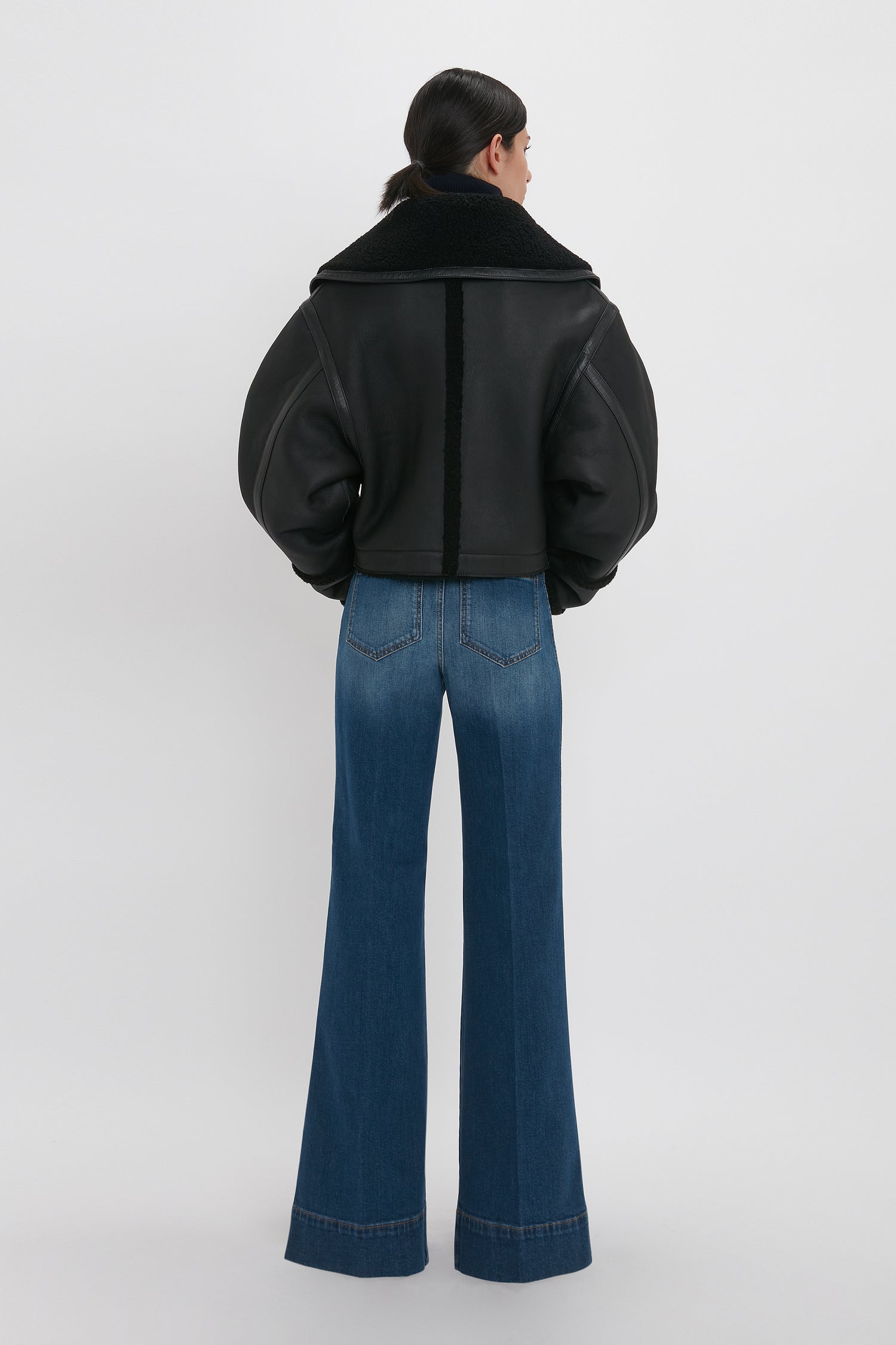 Person with long hair tied back, wearing a black Shearling Jacket In Black by Victoria Beckham and blue wide-leg jeans, standing and facing away.