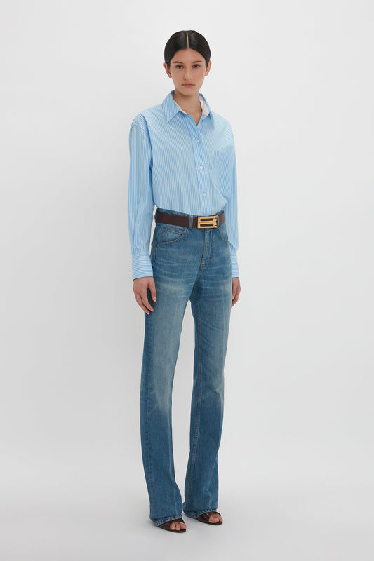 Person in a Cropped Long Sleeve Shirt In Marina-White by Victoria Beckham, blue jeans, brown belt, and black sandals standing against a plain white background.