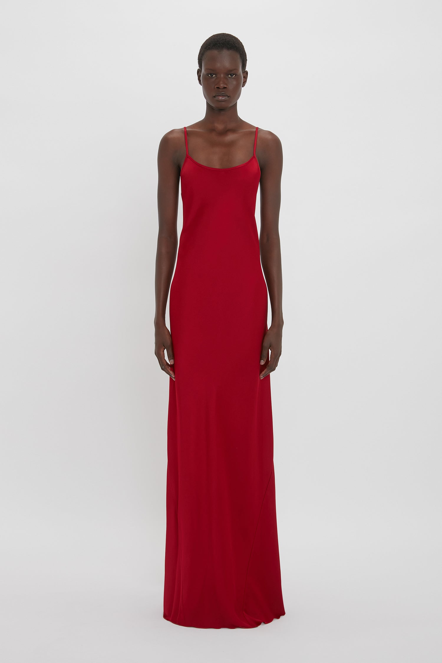 A person stands against a plain white background wearing a long, fitted Low Back Cami Floor-Length Dress In Poppy Red by Victoria Beckham with thin straps, reminiscent of a 1990s-inspired camisole-slip dress.