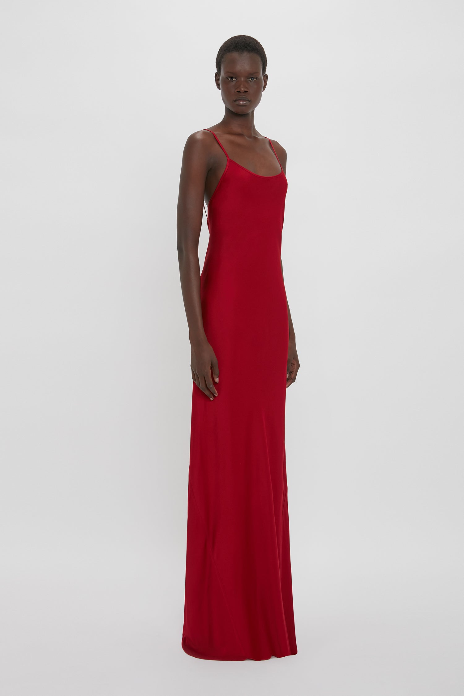 Person wearing a long, sleeveless 1990s-inspired **Victoria Beckham Low Back Cami Floor-Length Dress In Poppy Red** with thin straps, standing against a plain white background.