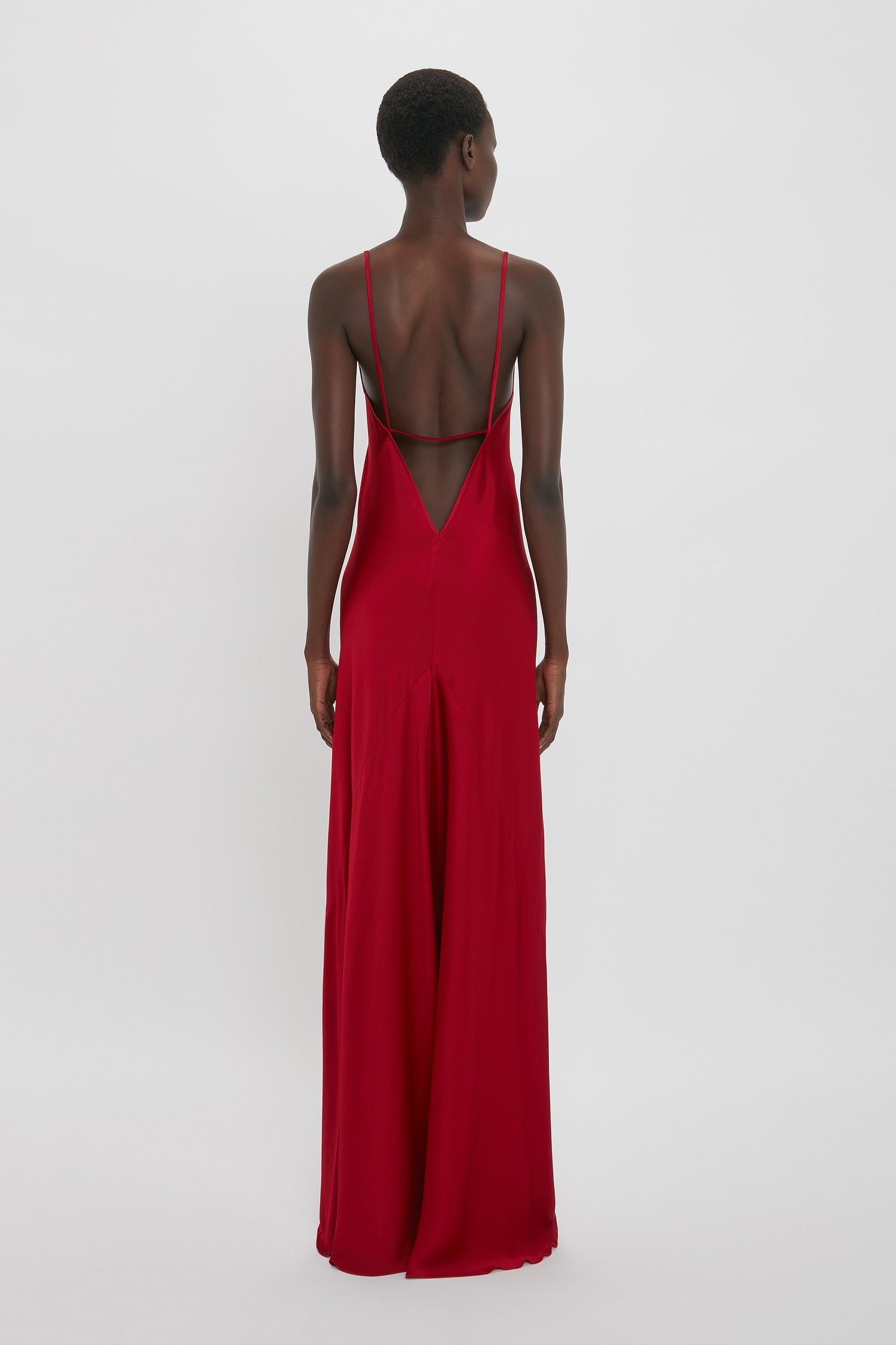 A person is standing facing away, wearing a poppy red, sleeveless long dress with thin shoulder straps and an open back against a plain white background. The Victoria Beckham Low Back Cami Floor-Length Dress In Poppy Red has a bias cut that drapes beautifully.