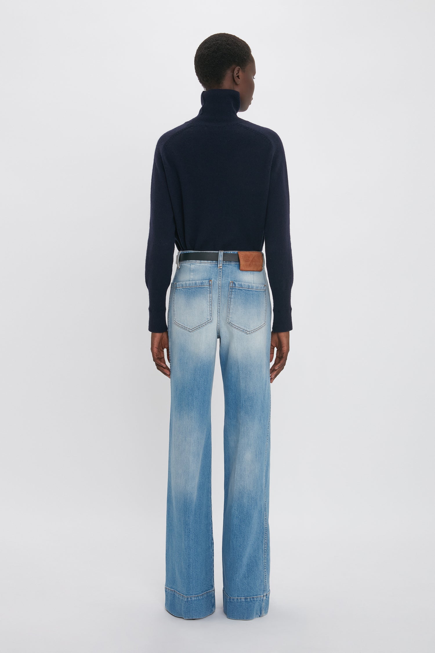 Person standing with back to the camera, wearing a navy turtleneck sweater and Alina High Waisted Jean In Light Summer Wash by Victoria Beckham reminiscent of vintage denim style.