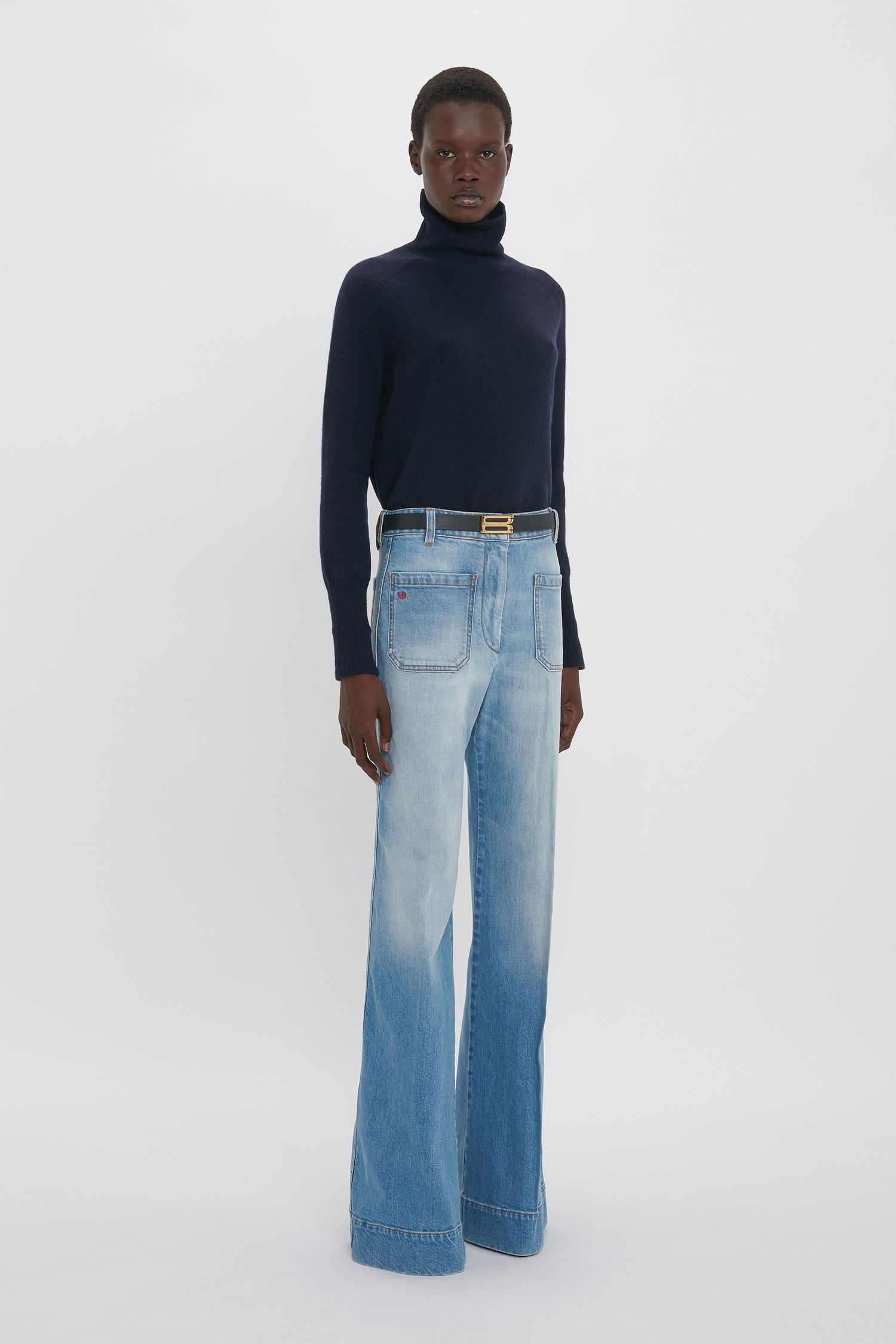 A person stands against a plain background wearing a black turtleneck sweater, Alina High Waisted Jean In Light Summer Wash by Victoria Beckham, and a belt.