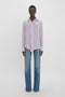 Person wearing an ethereal shade of Victoria Beckham Asymmetric Ruffle Blouse In Petunia paired with wide-leg blue jeans, standing against a plain white background.