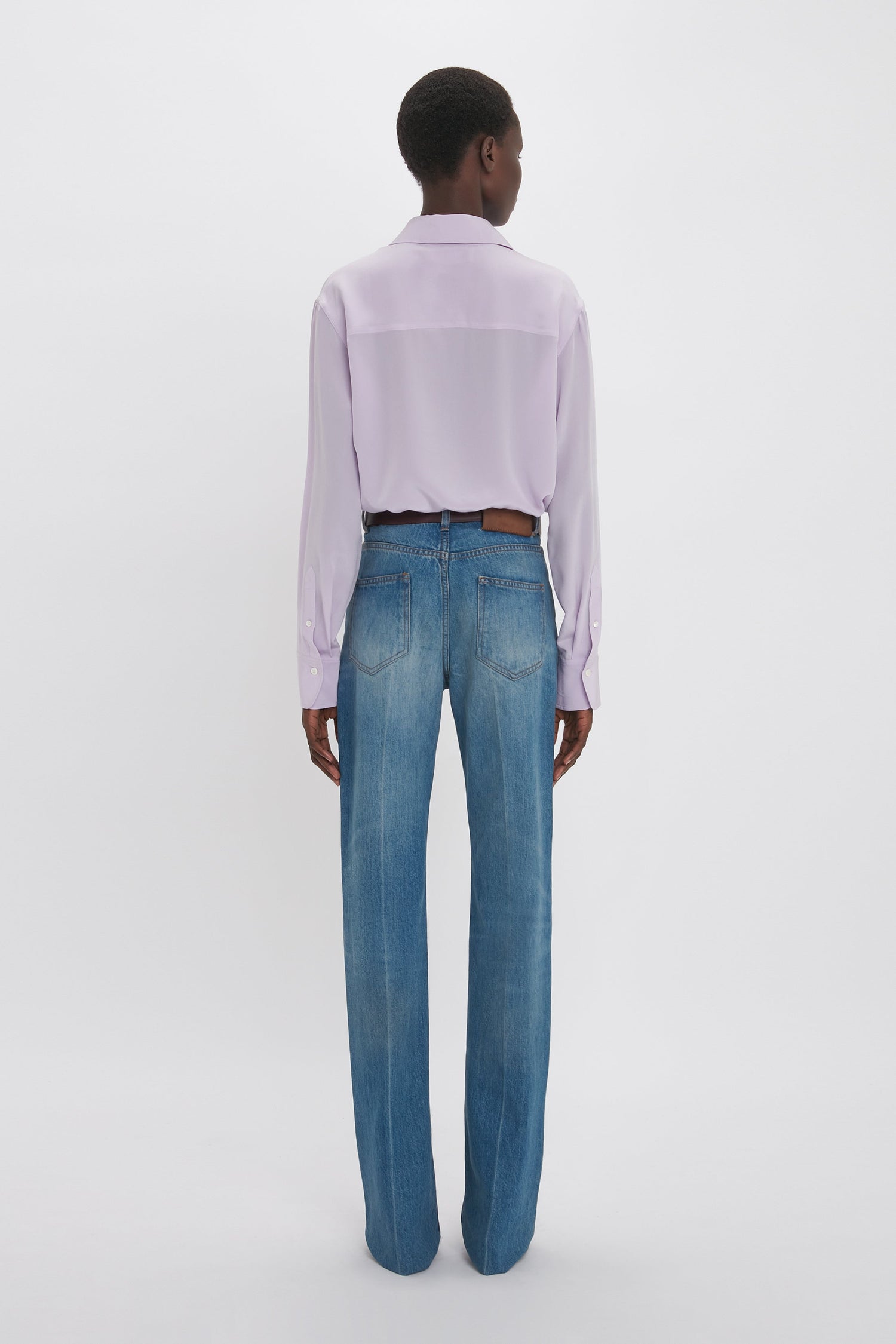 Rear view of a person wearing a Victoria Beckham asymmetric ruffle blouse in petunia and blue jeans, standing against a white background.