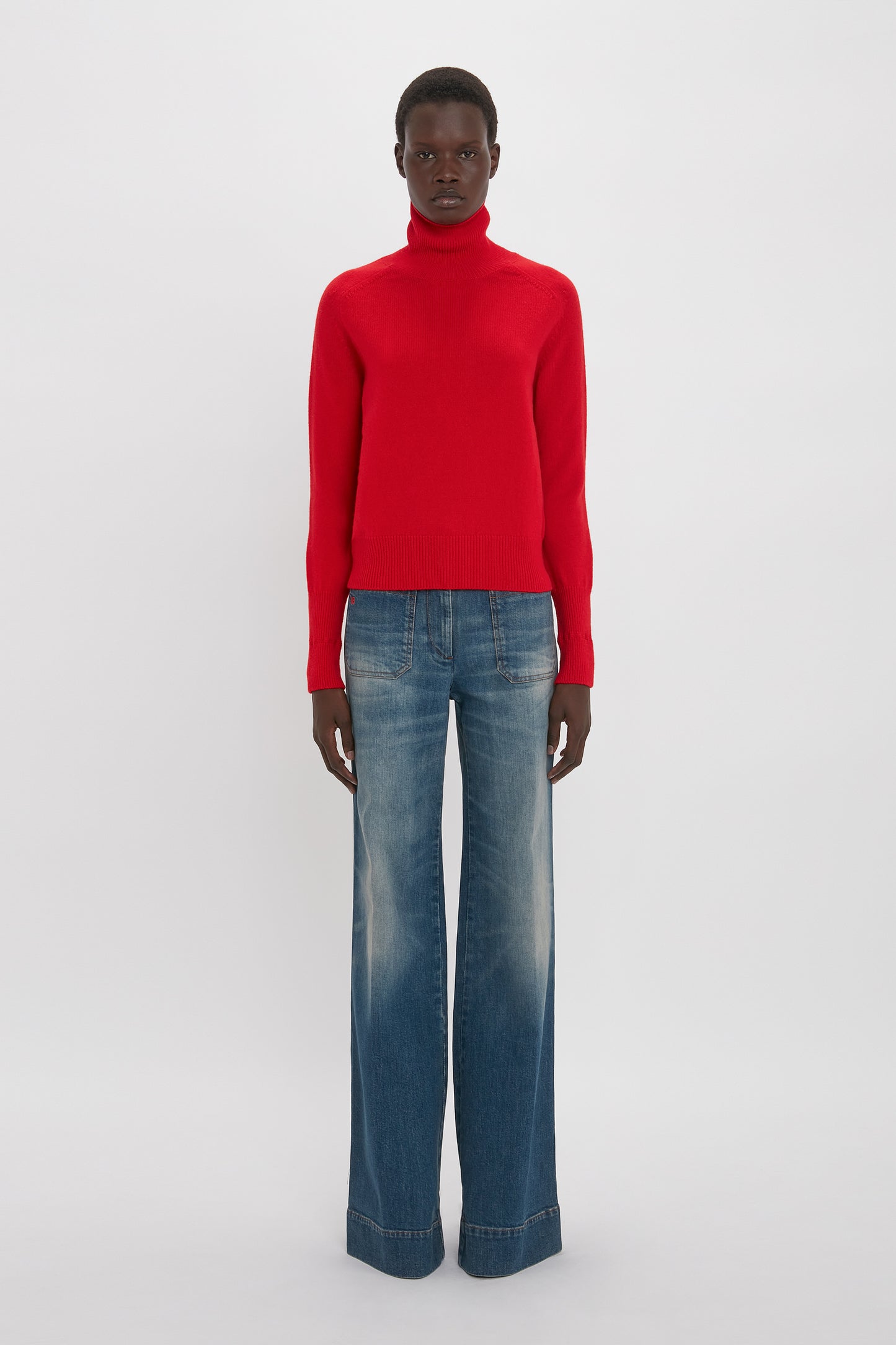 A person stands against a plain backdrop wearing a red turtleneck sweater and Victoria Beckham's Alina High Waisted Jean In Heavy Vintage Indigo Wash. They face forward with a neutral expression, evoking a touch of Victoria Beckham's sleek style.