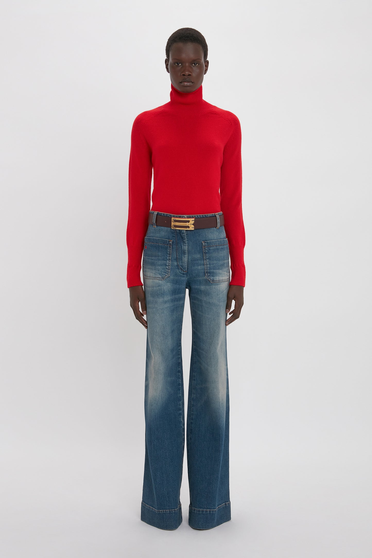 Person wearing a red turtleneck sweater and Victoria Beckham Alina High Waisted Jean In Heavy Vintage Indigo Wash, standing against a plain white background.