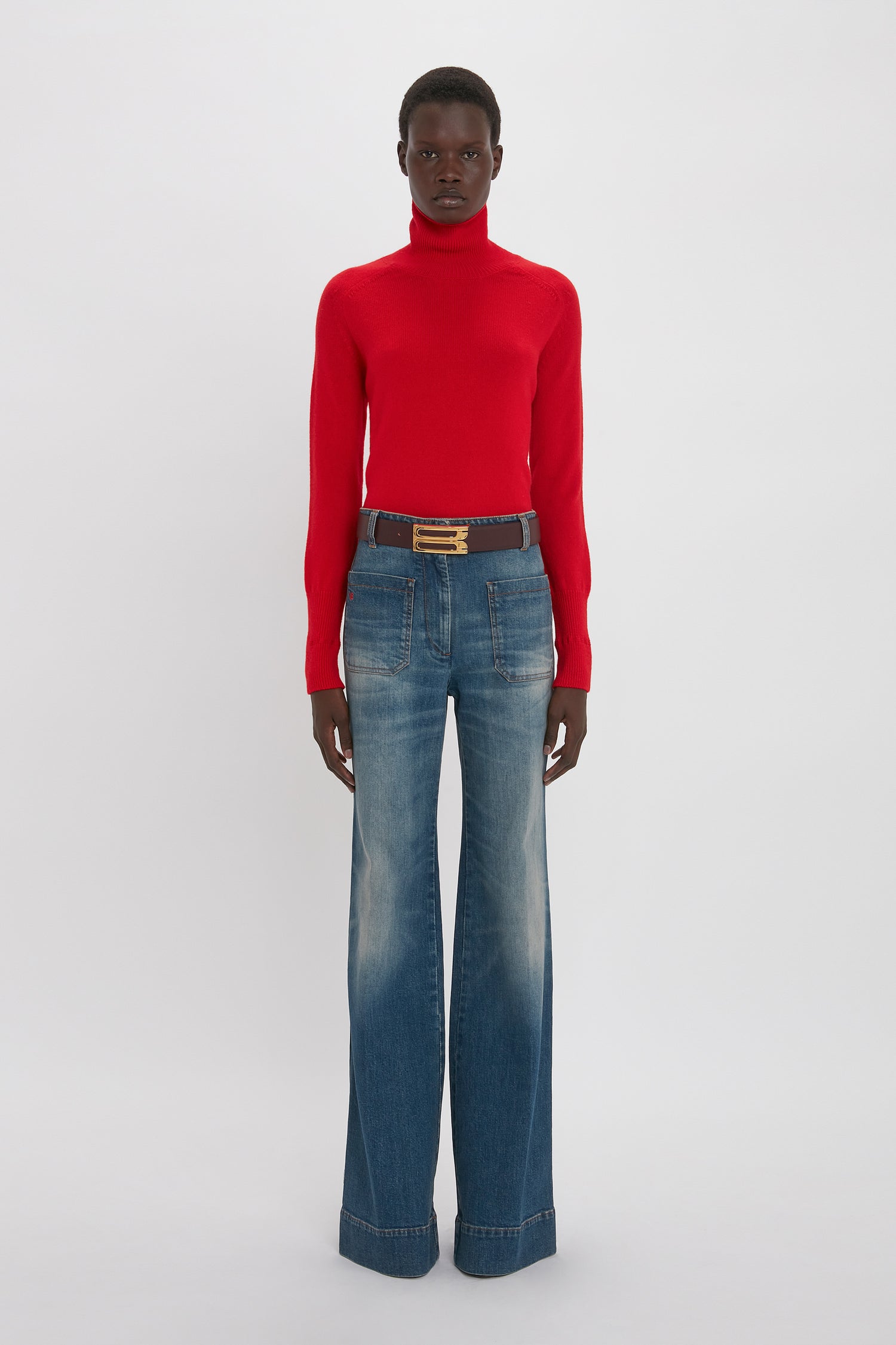 Person wearing a red turtleneck sweater and Victoria Beckham Alina High Waisted Jean In Heavy Vintage Indigo Wash, standing against a plain white background.