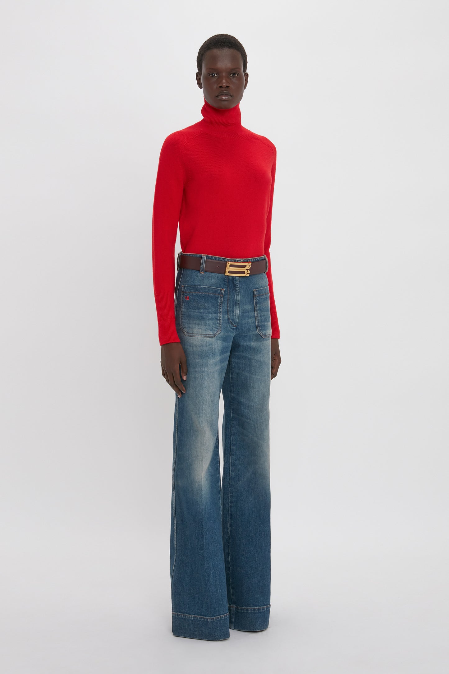 A person stands against a white background wearing a red turtleneck sweater, Alina High Waisted Jean In Heavy Vintage Indigo Wash by Victoria Beckham, and a black belt with a gold buckle.