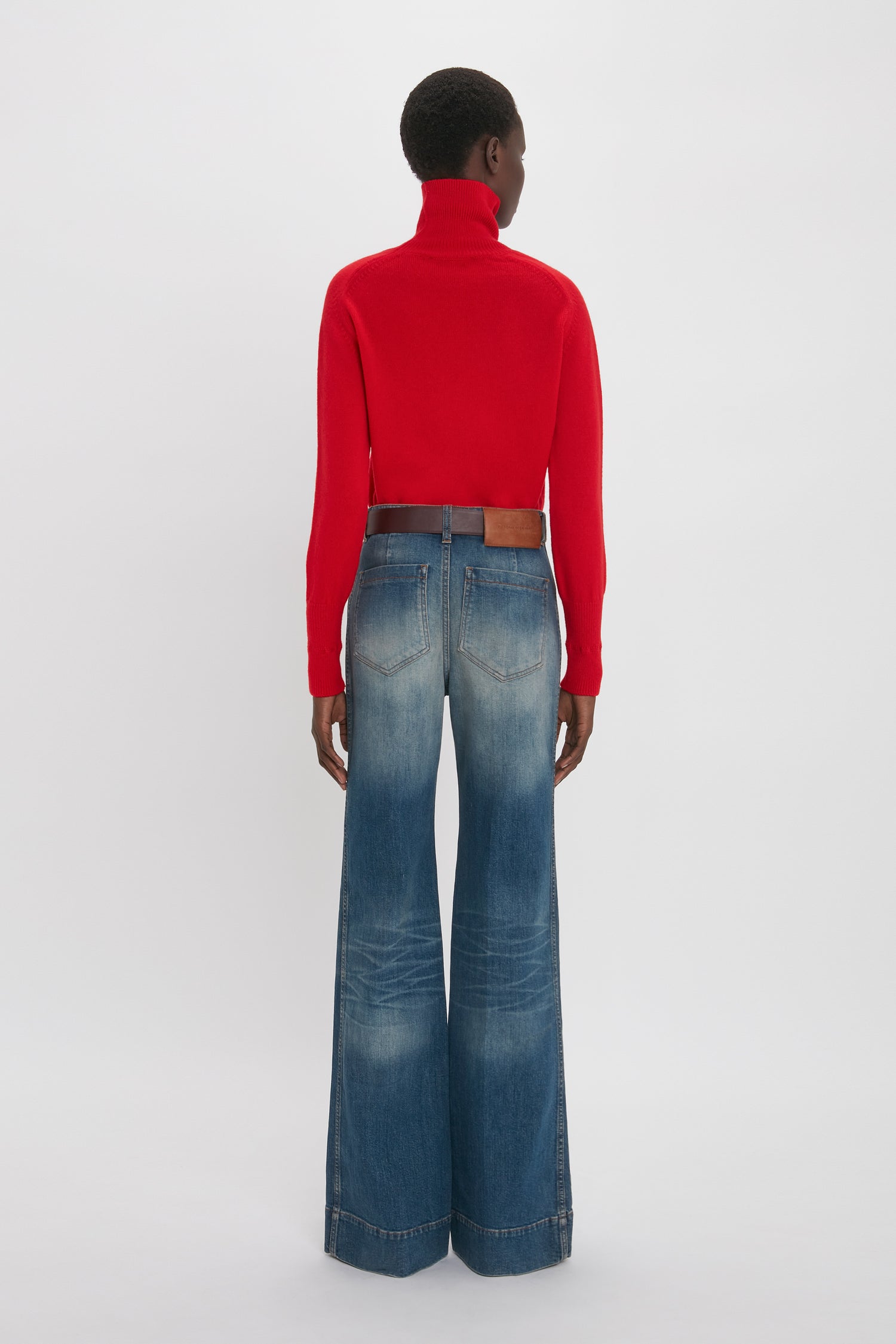 Person seen from the back, wearing a red turtleneck sweater and Alina High Waisted Jean In Heavy Vintage Indigo Wash with a brown belt, reminiscent of Victoria Beckham's iconic style.