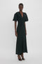 Person standing and modeling a Gathered V-Neck Midi Dress in Black by Victoria Beckham with short puffed sleeves and a gathered detail at the waist, crafted from figure-flattering stretch fabric for a contemporary edge. The dress is paired with black high-heeled shoes.