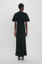 A person stands facing away from the camera, wearing a Victoria Beckham Gathered V-Neck Midi Dress in Black, short-sleeved and crafted from figure-flattering stretch fabric, paired with black high-heeled shoes against a plain white background.