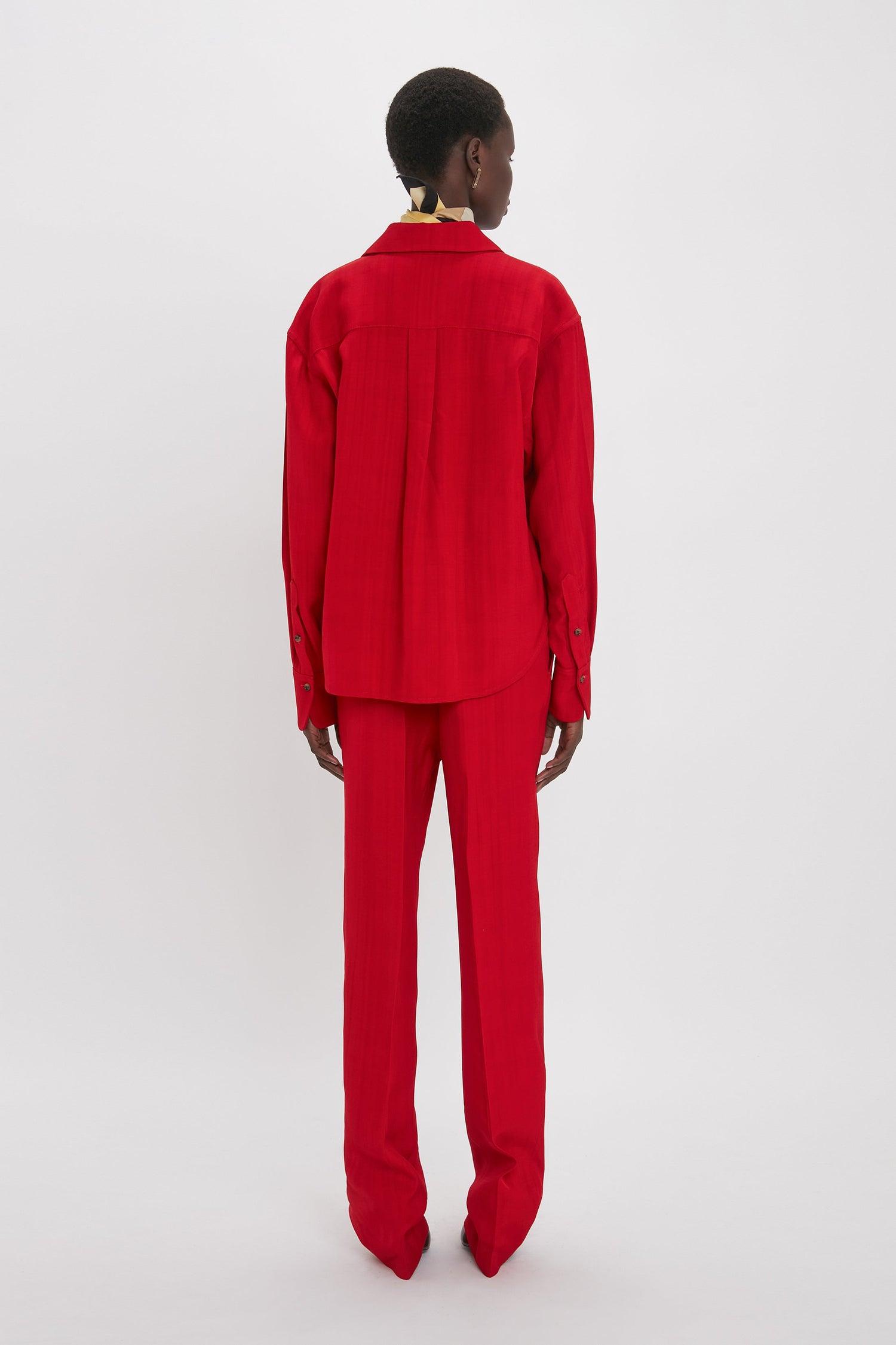 A person stands facing away, showcasing a lean silhouette in a long-sleeved red shirt and matching Victoria Beckham Tapered Leg Trouser In Carmine against a plain white background.