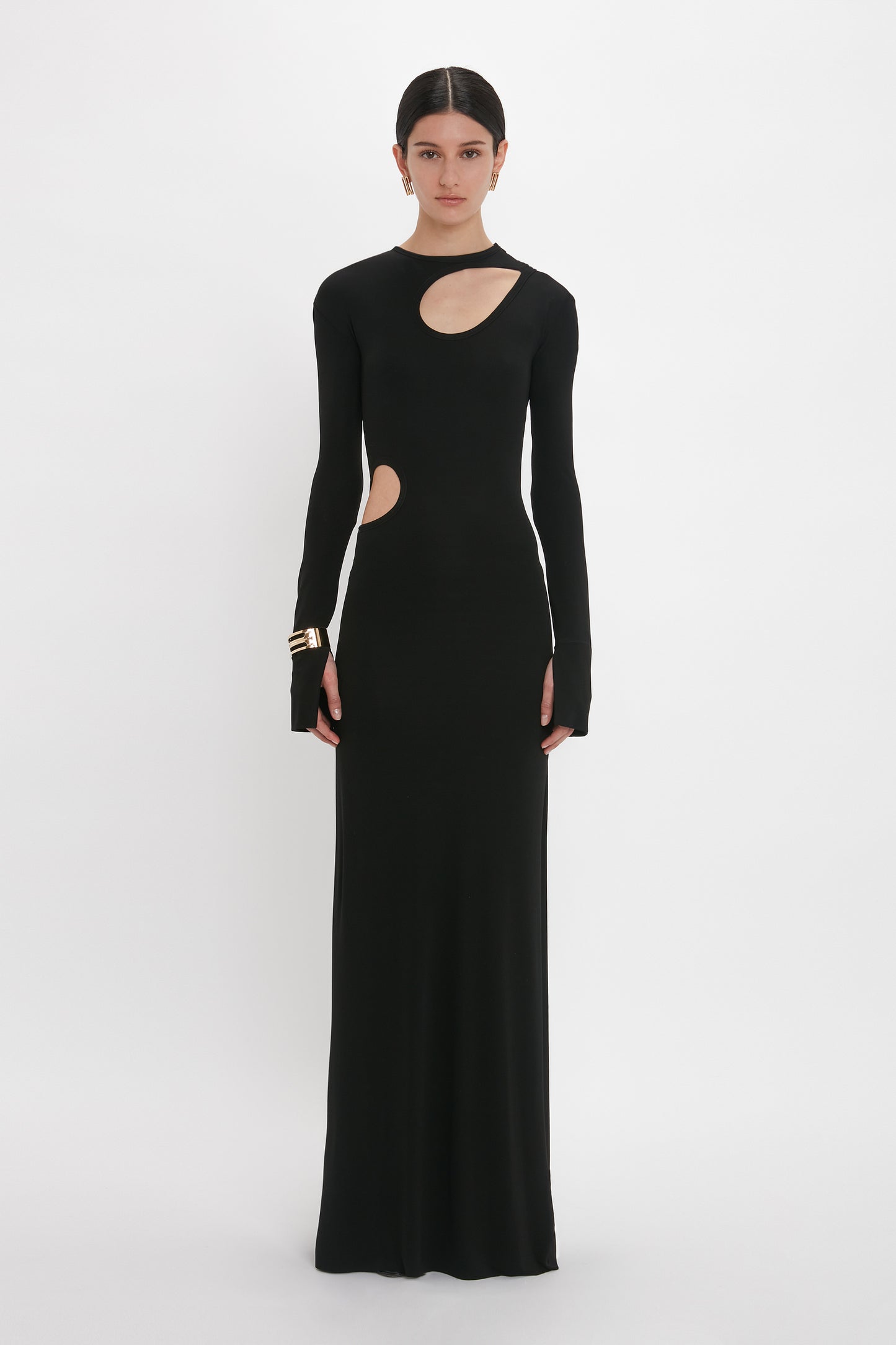 A person is standing against a plain white background wearing a long black Cut-Out Jersey Floor-Length Dress In Black with shoulder cutouts and a side cutout, channeling the elegant style of Victoria Beckham. Gold bracelets add a touch of sophistication to the evening gown.