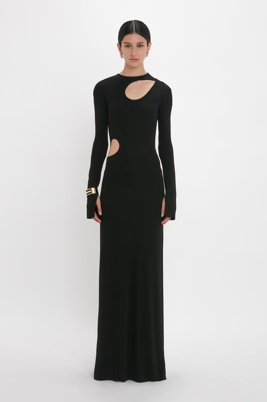 A person is standing against a plain white background wearing a long black Cut-Out Jersey Floor-Length Dress In Black with shoulder cutouts and a side cutout, channeling the elegant style of Victoria Beckham. Gold bracelets add a touch of sophistication to the evening gown.