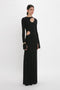 A person stands wearing a long, fitted Cut-Out Jersey Floor-Length Dress In Black by Victoria Beckham. The evening gown features cut-out details at the chest and waist, complemented by long sleeves and a high neckline. The person is looking slightly to the side.