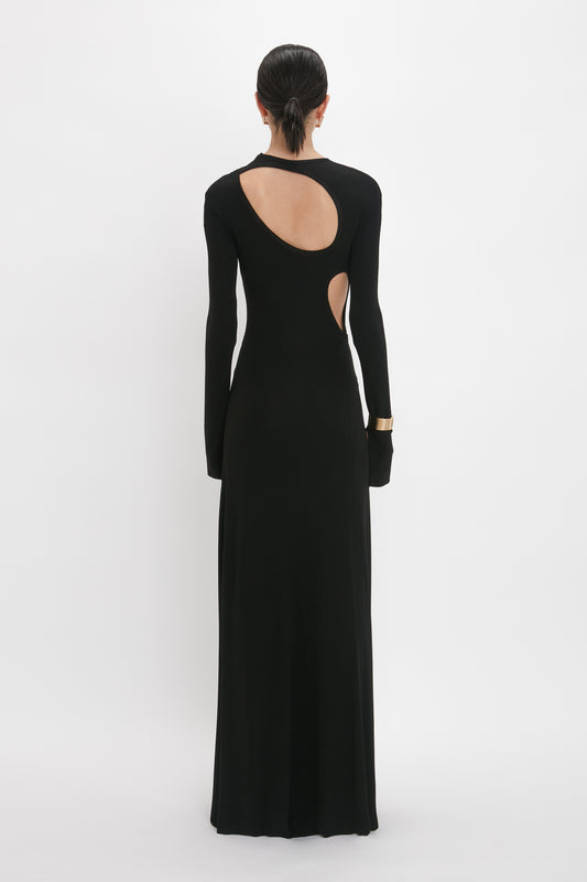 A person is standing with their back to the camera, wearing a Victoria Beckham Cut-Out Jersey Floor-Length Dress In Black. Their hair is styled back, and they are wearing a metallic cuff bracelet.