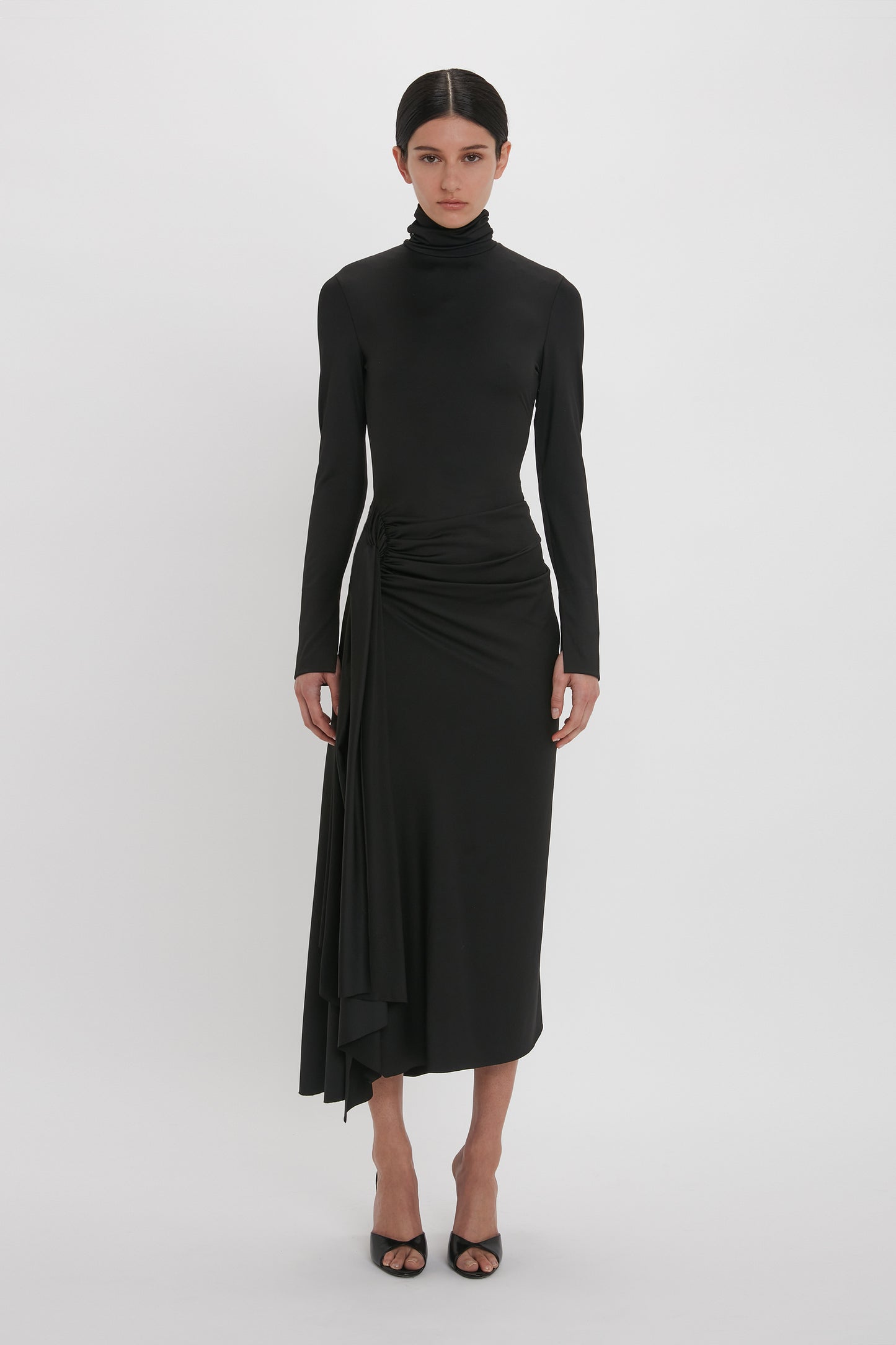 A woman in a sleek black Victoria Beckham gown with a high neckline, posing against a plain white background.