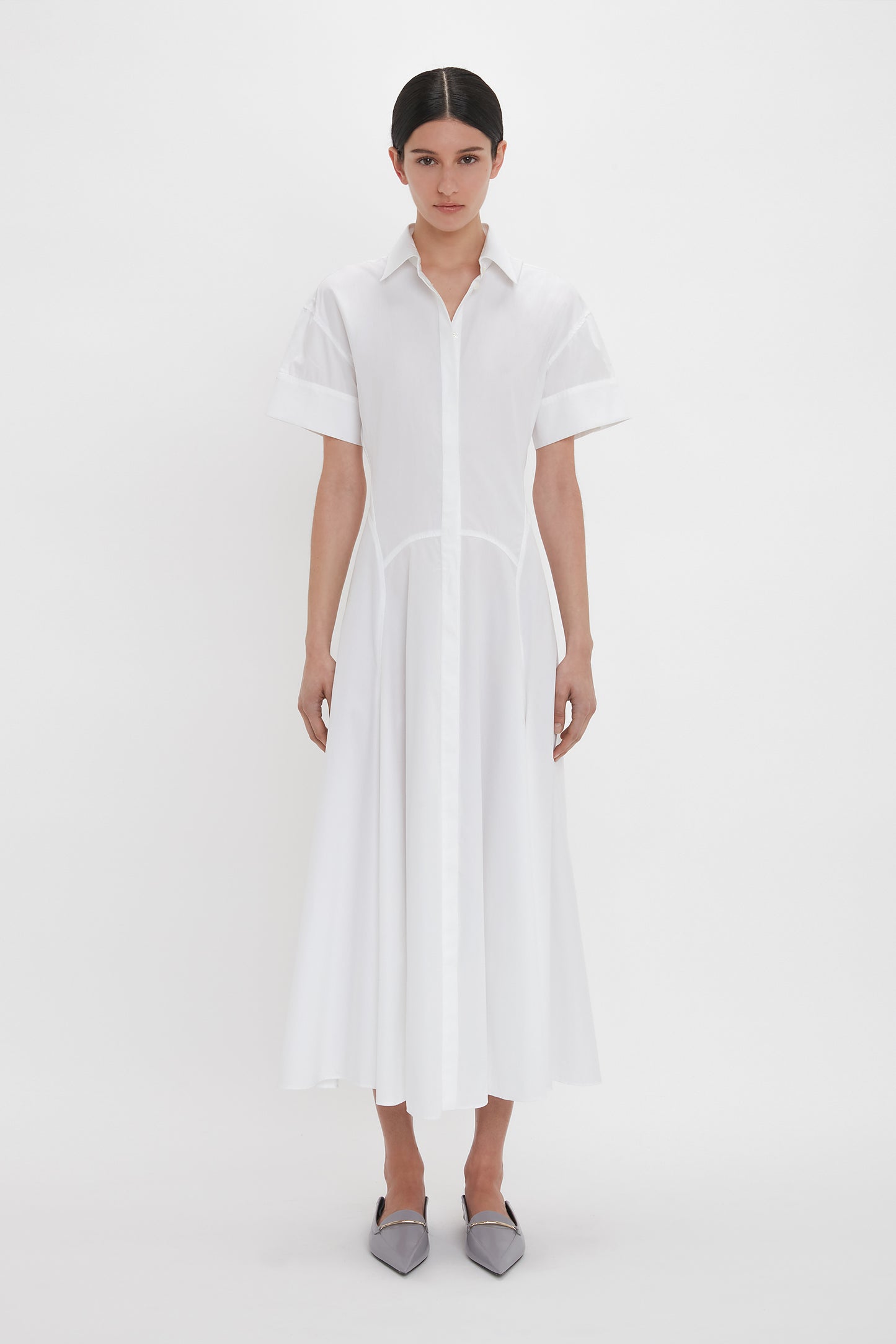 A person stands against a white background wearing a mid-length Panelled Shirt Dress In White by Victoria Beckham made from organic cotton poplin, with short sleeves and grey pointed shoes.