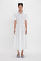 A person stands against a white background wearing a mid-length Panelled Shirt Dress In White by Victoria Beckham made from organic cotton poplin, with short sleeves and grey pointed shoes.