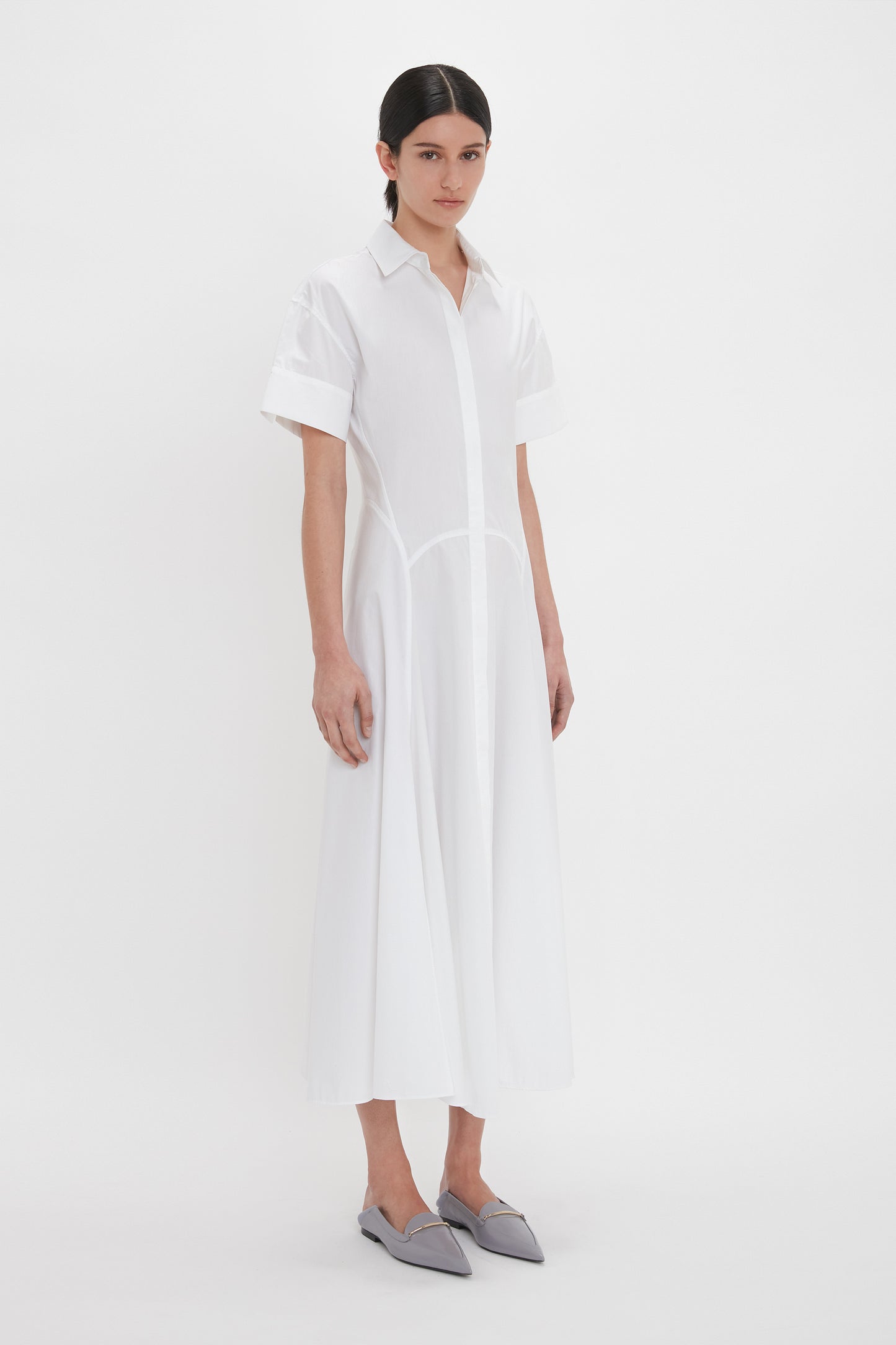 A person stands against a white background wearing the Panelled Shirt Dress In White from Victoria Beckham and grey pointed-toe flats. They have dark hair pulled back neatly.