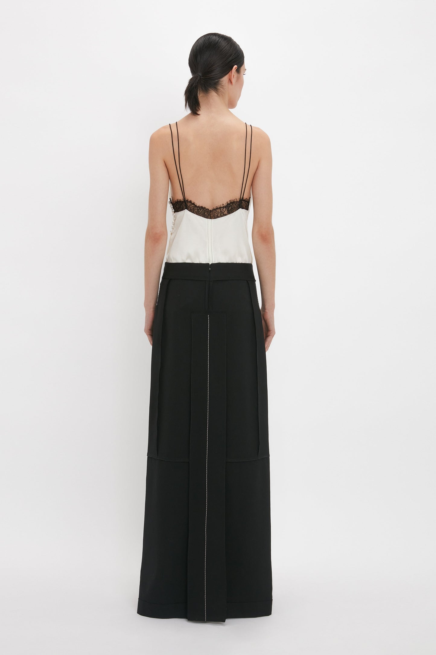 A person stands with their back to the camera, wearing a Victoria Beckham Lace Detail Cami Top In Harvest Ivory and a long black skirt with visible seams, on a plain white background.