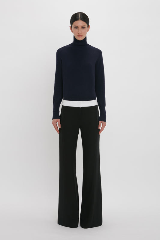 A person with dark hair wears a black turtleneck sweater and Victoria Beckham Side Panel Trouser In Black, standing against a plain white background.