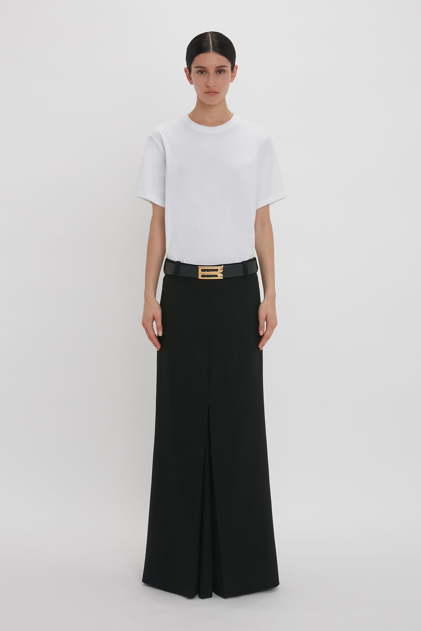 A woman stands wearing a plain white t-shirt and a Victoria Beckham floor-length box pleat skirt in black with a black belt that has a gold buckle.