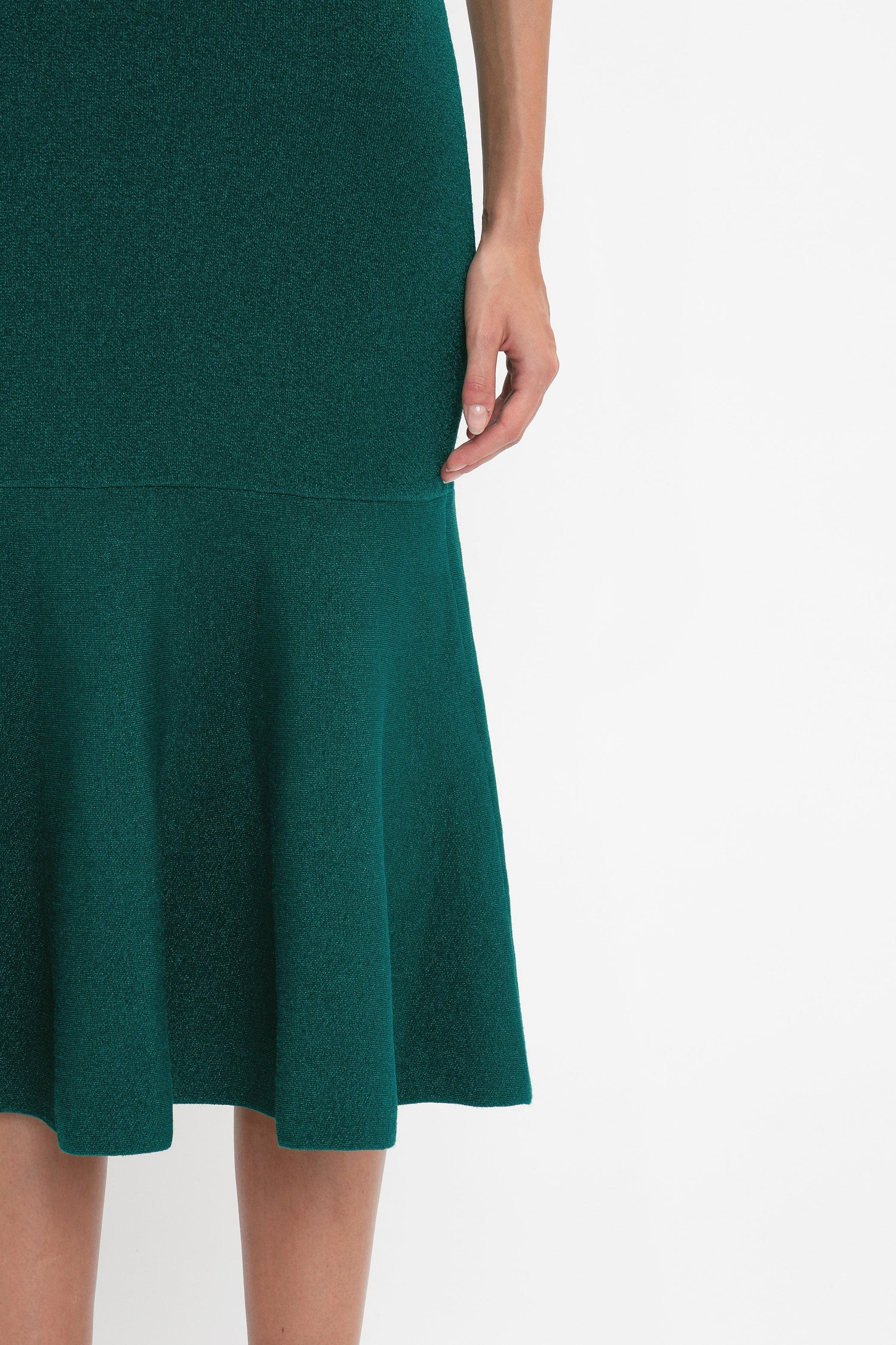 A person wearing a fitted Victoria Beckham VB Body Flared Skirt In Lurex Green with a flared silhouette hem. The image shows the lower half of their body, with one arm resting by their side.